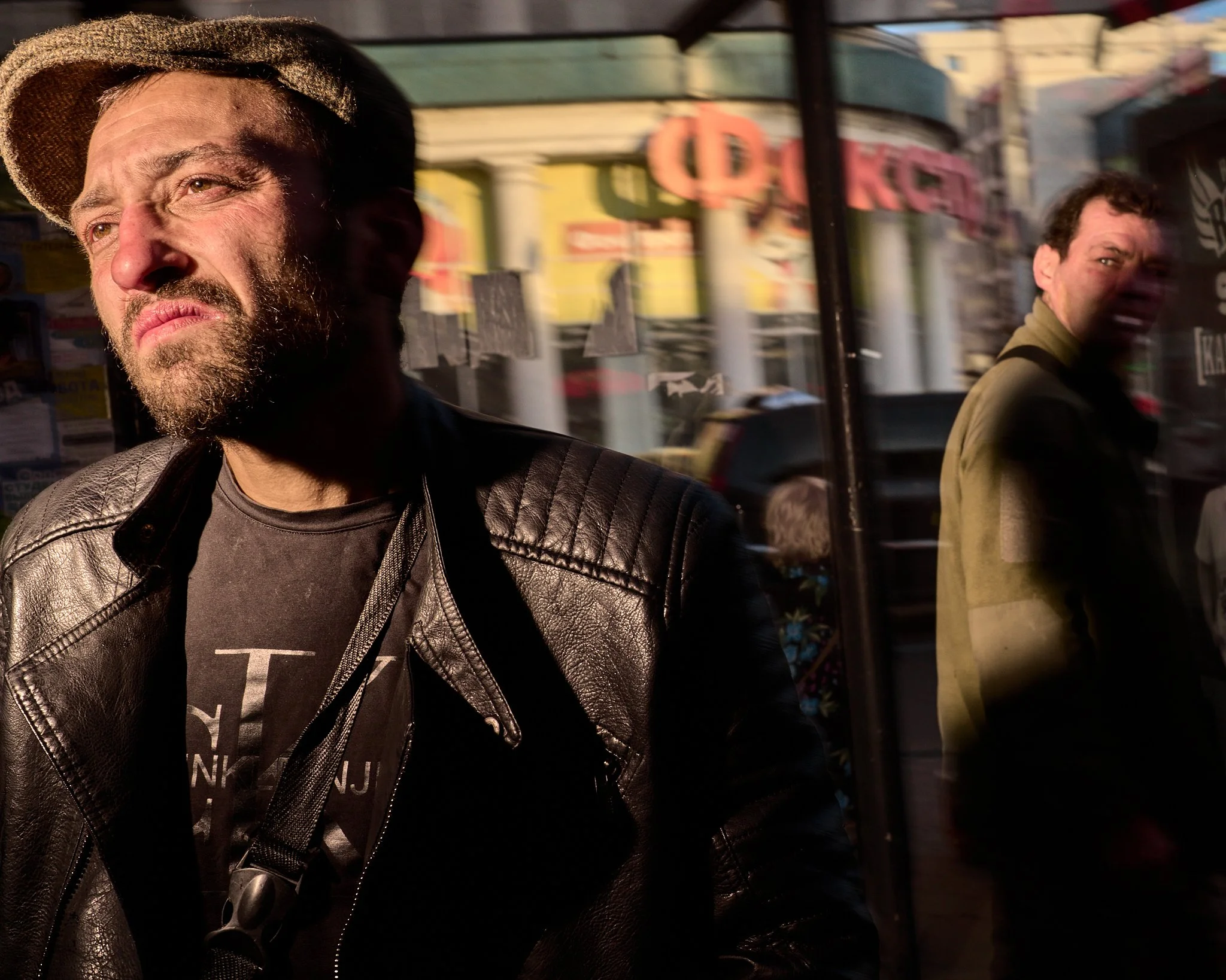 Two men standing outside a glass storefront. The man in the foreground is wearing a leather jacket and a flat cap, squinting into the sunlight. The reflection and background show colorful city buildings and people passing by.