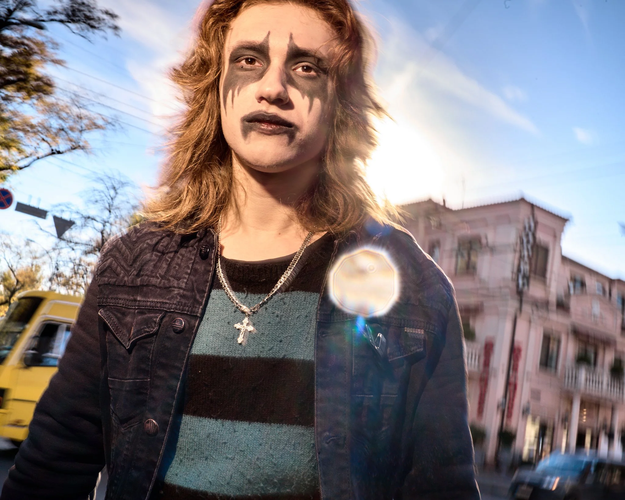 Young man with long hair and face makeup resembling heavy metal or gothic style, wearing a black denim jacket, striped shirt, and a cross necklace, standing outdoors with sunlight behind him, near a pink building and a yellow truck.