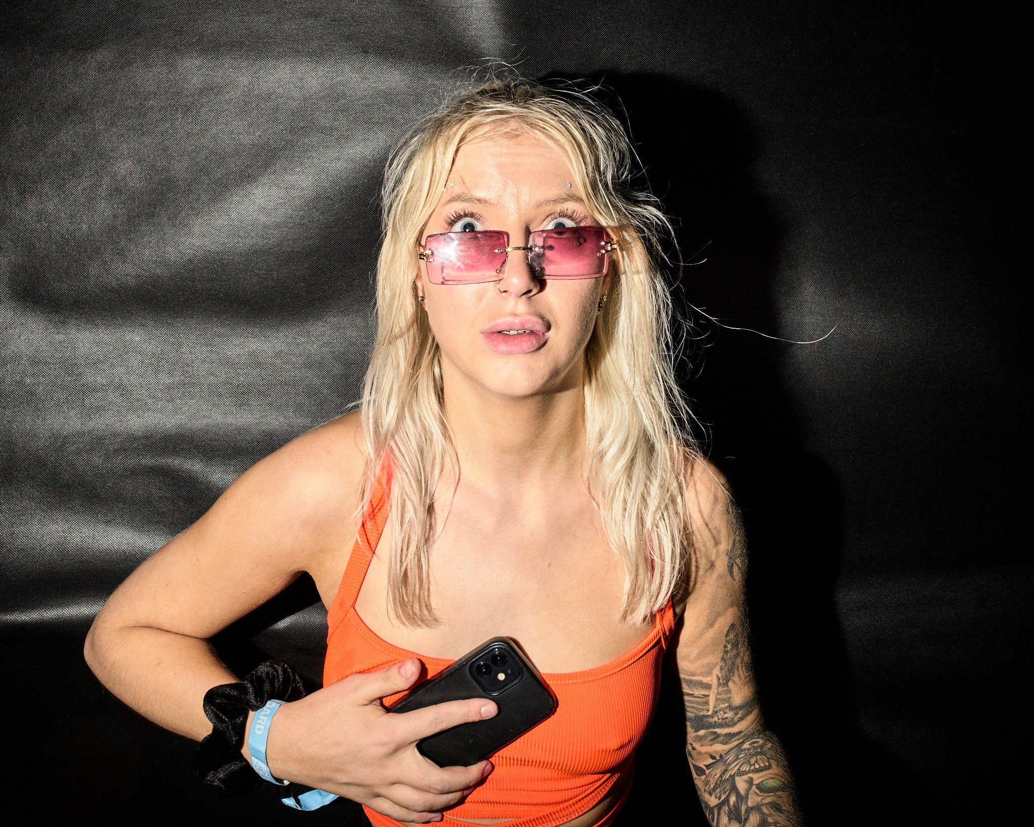 A woman with long blonde hair, wearing pink sunglasses, an orange tank top, holding a phone, and displaying a surprised or confused expression. She has tattoos on her arm and is standing against a black background.