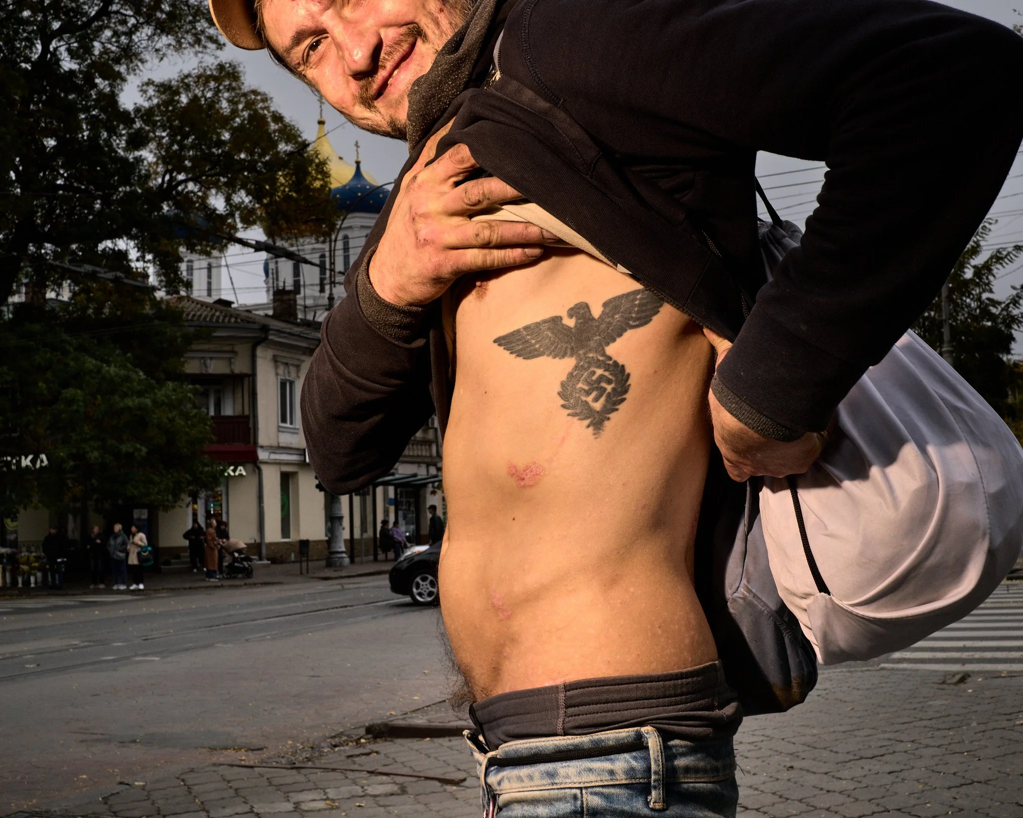 A person is holding up their black jacket to reveal a large Nazi eagle tattoo with a swastika on their chest.