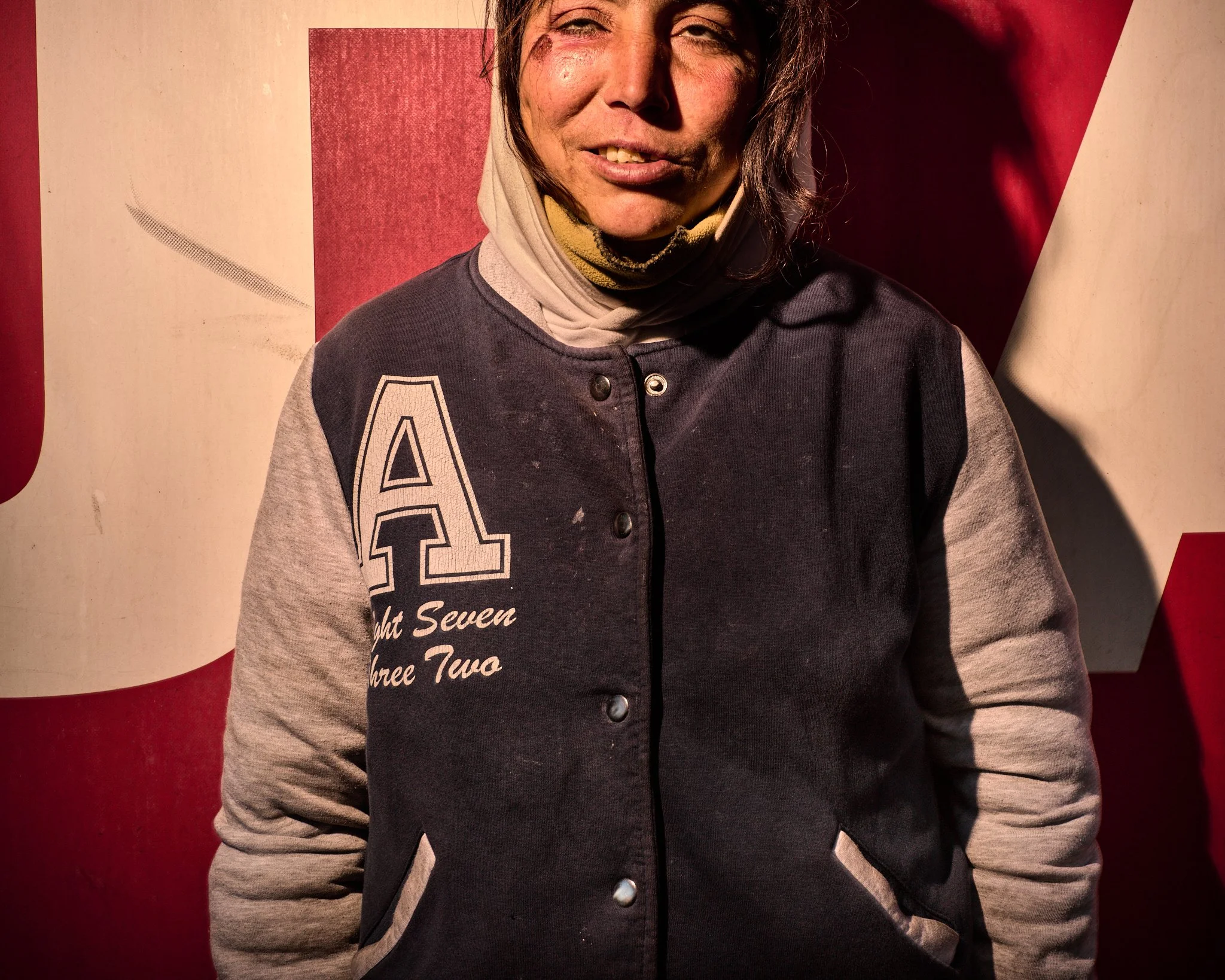 A young woman with a smirk, wearing a black and gray jacket with a large letter 'A' on it, standing in front of a red and white background.