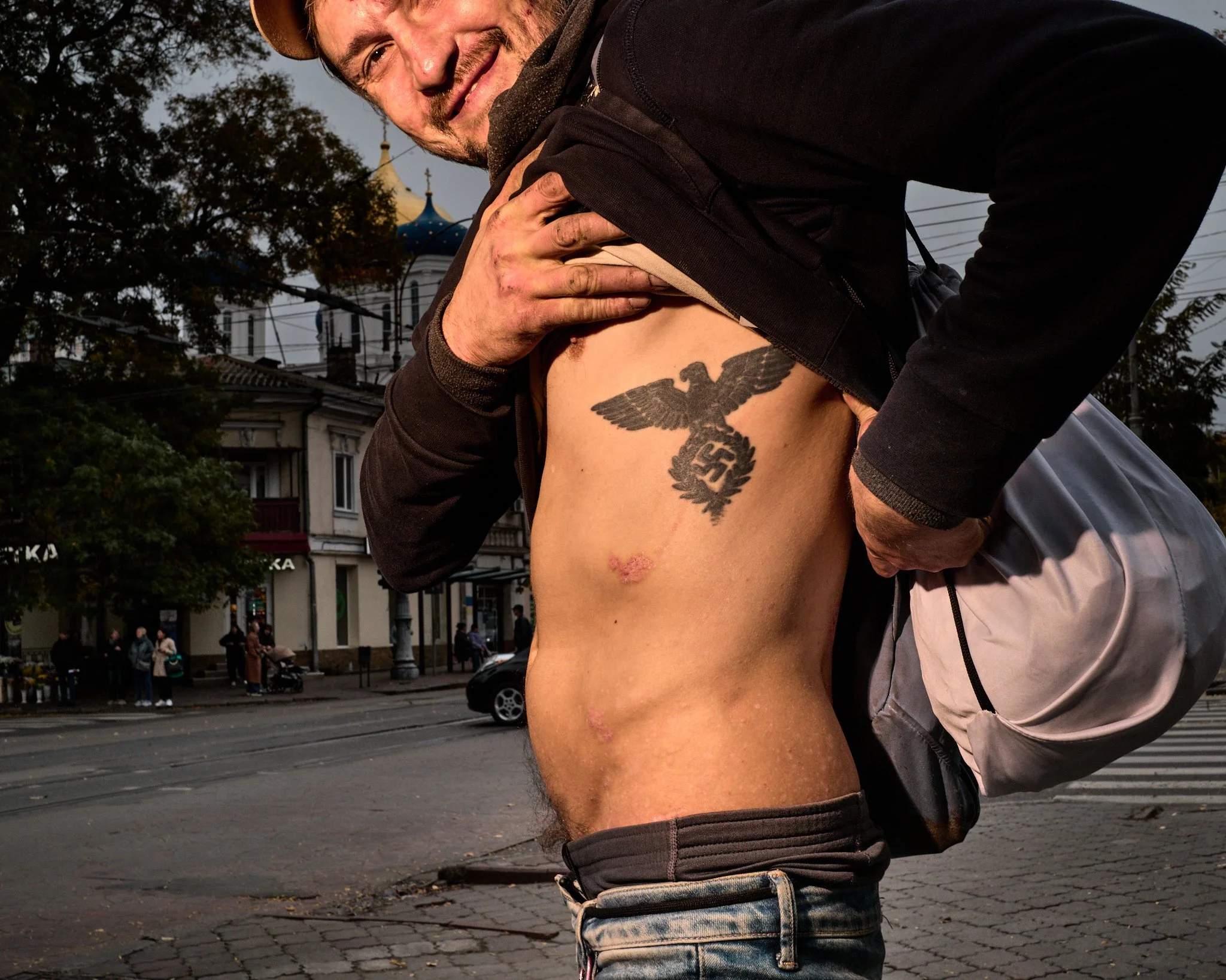 A smiling man lifts his shirt to reveal a Nazi tattoo on his side, with a swastika and eagle emblem, in an urban street setting during daylight.
