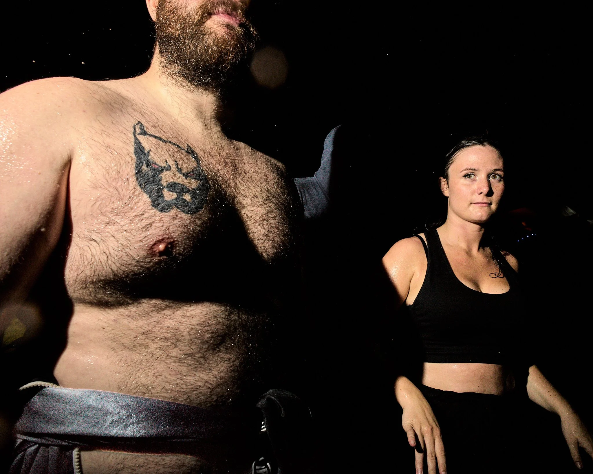 A shirtless man with a beard and chest hair, with a tattoo on his left chest, and a woman with dark hair in a ponytail wearing a black sports bra, standing in a dark environment with some lighting.