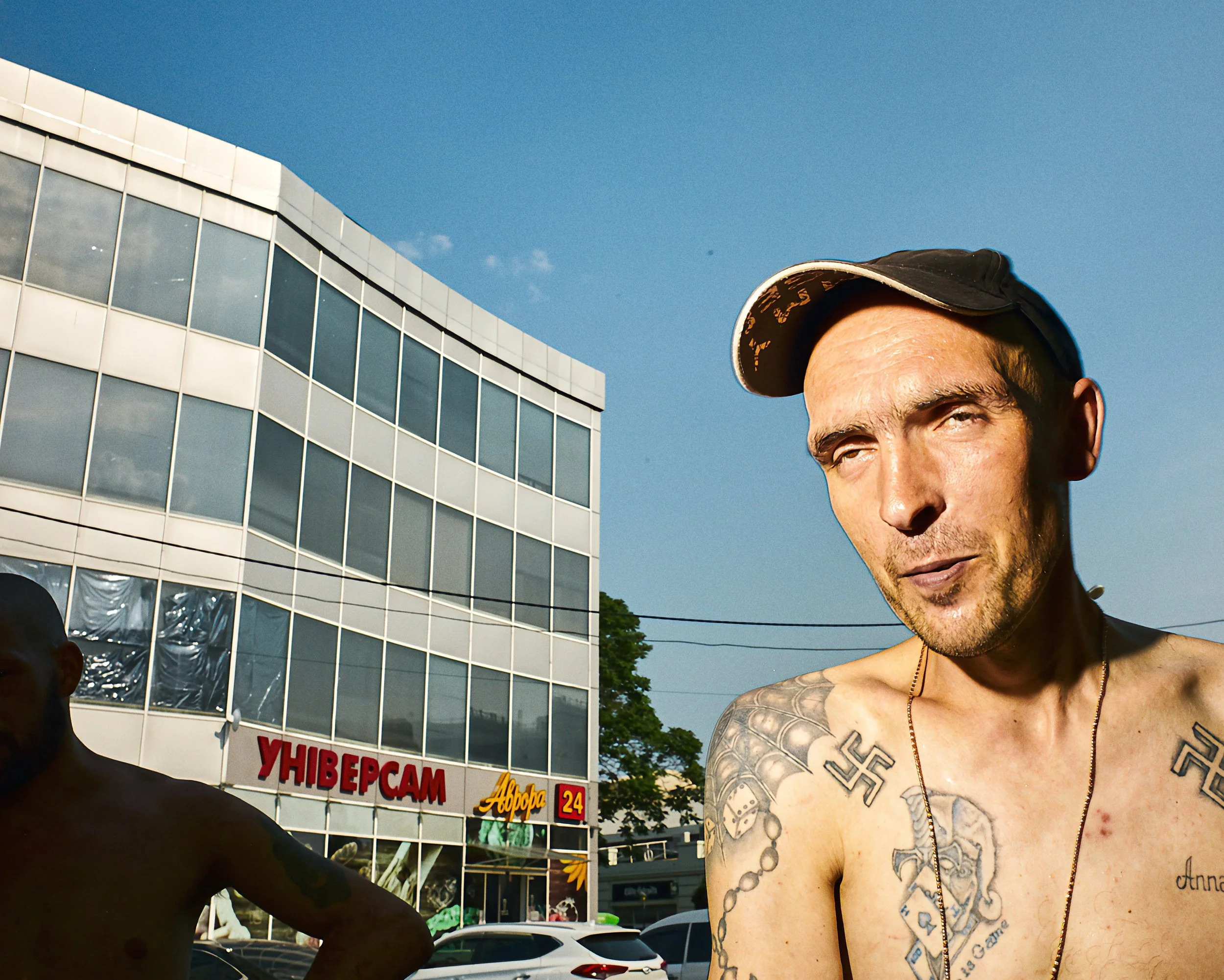 Close-up of a tattooed shirtless man wearing a black cap, with part of his face visible, standing outdoors with a building and parked cars in the background on a sunny day.