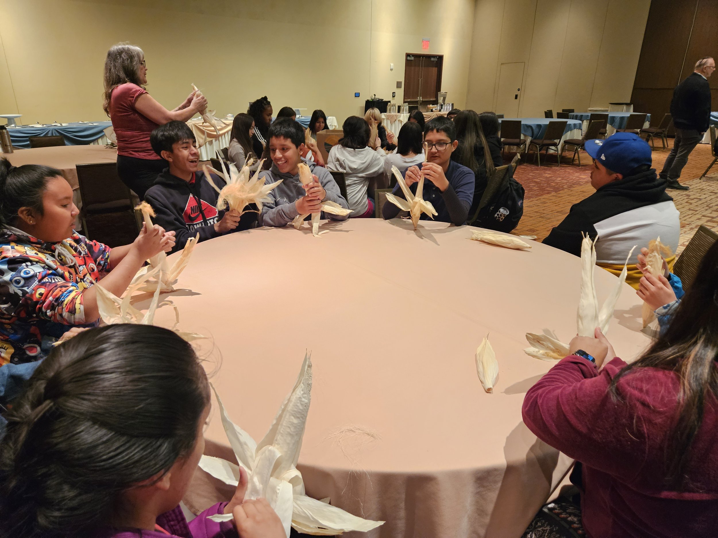 A group of children sitting around a circular table holding corn husks, possibly preparing food or participating in a craft activity, with a woman in the background.