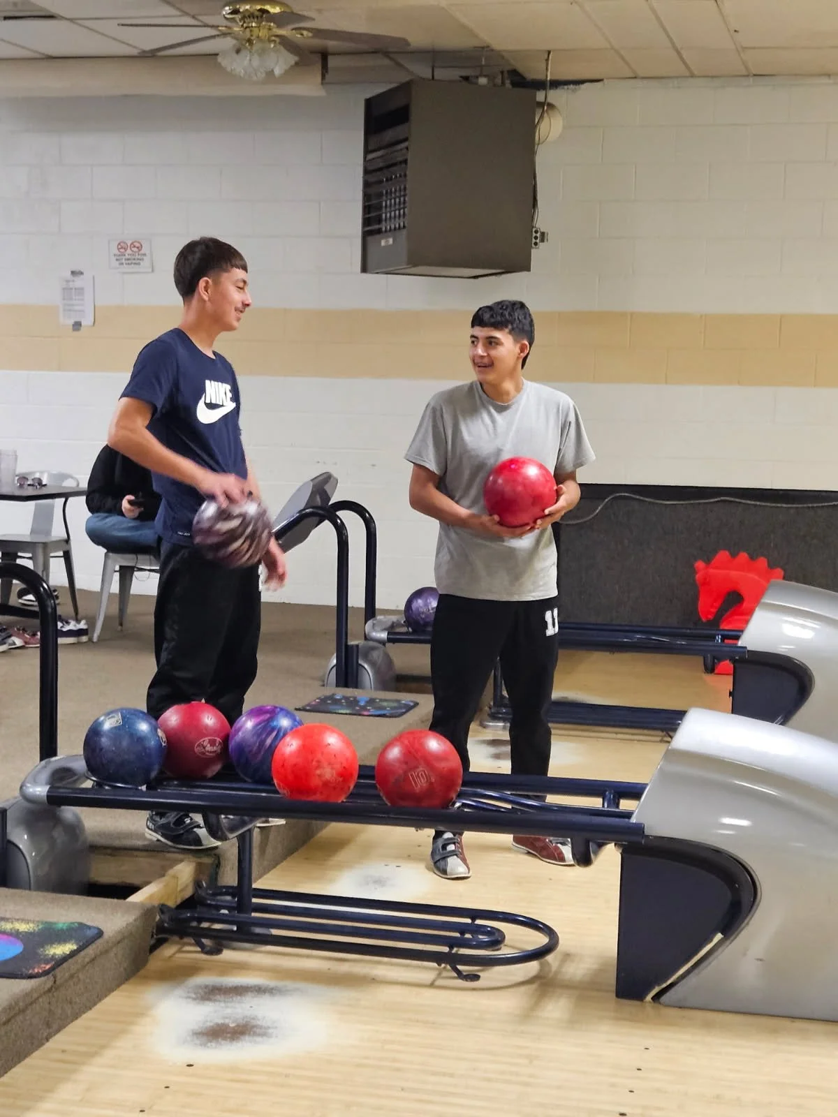 Two young men standing at a bowling alley, one holding a red bowling ball, smiling, the other holding a black and white ball, engaging in conversation. Several other bowling balls on a rack in the foreground, with a floor of wood and a beige brick wa