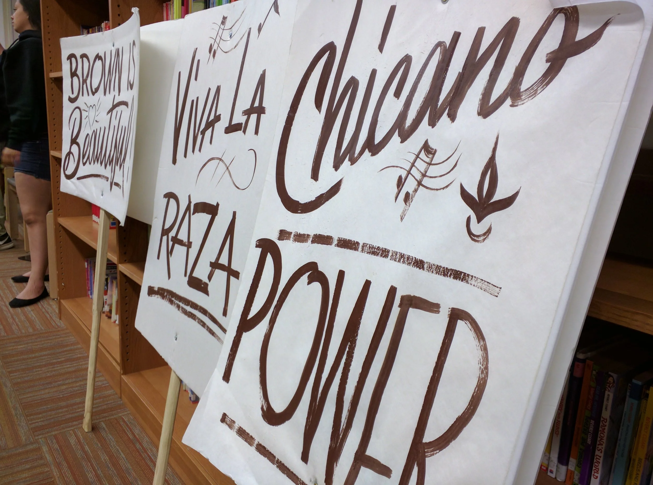 Handwritten signs on large sheets of paper displayed on stands. The signs have Spanish phrases including 'BROWNS BEAUTIFUL,' 'VIVA LA IGNANCIA,' and 'RAZA POR LA.'