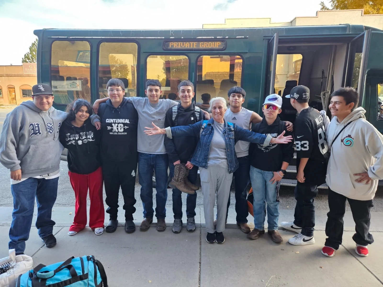 Group of teenagers and an adult woman standing in front of a school bus with a sign 'Private Group', smiling with arms around each other.