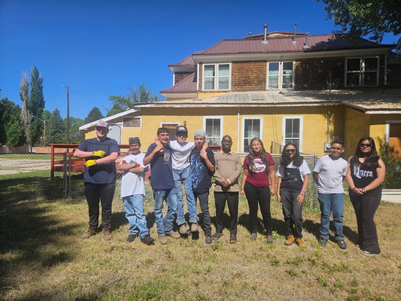 A group of nine diverse young people standing on grass in front of a yellow house under a blue sky.