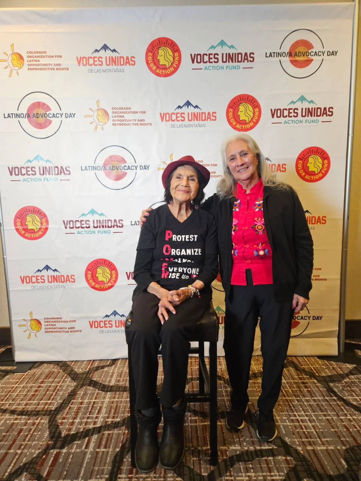 Two women standing in front of a backdrop with logos and text celebrating Latino Advocacy Day. One woman is seated on a stool, wearing black pants, a black shirt with the acronym 'PROTEST ORGANIZE EVERYONE FIGHT' in colorful letters, and a red hat. T