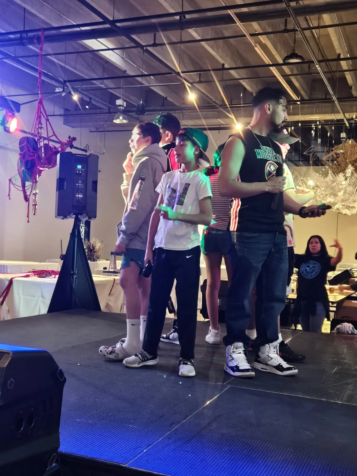 Group of young people on a stage at a nightclub or event, some holding microphones and a phone, with colorful lighting and a speaker indoor setting.