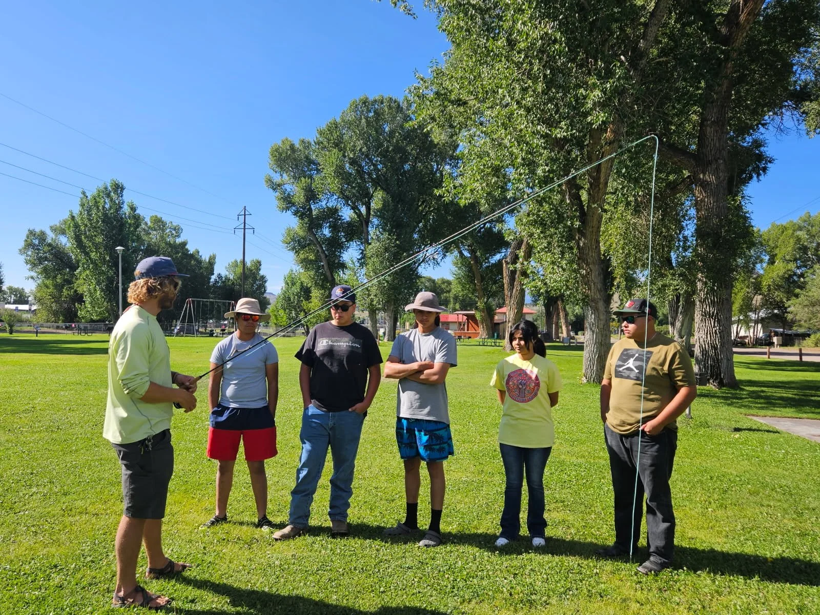 Group of six people standing outdoors on grass, with five listening to a man holding a fishing pole. The group includes both men and women wearing casual summer clothing, and some are wearing hats and sunglasses. There are trees, a playground, and cl