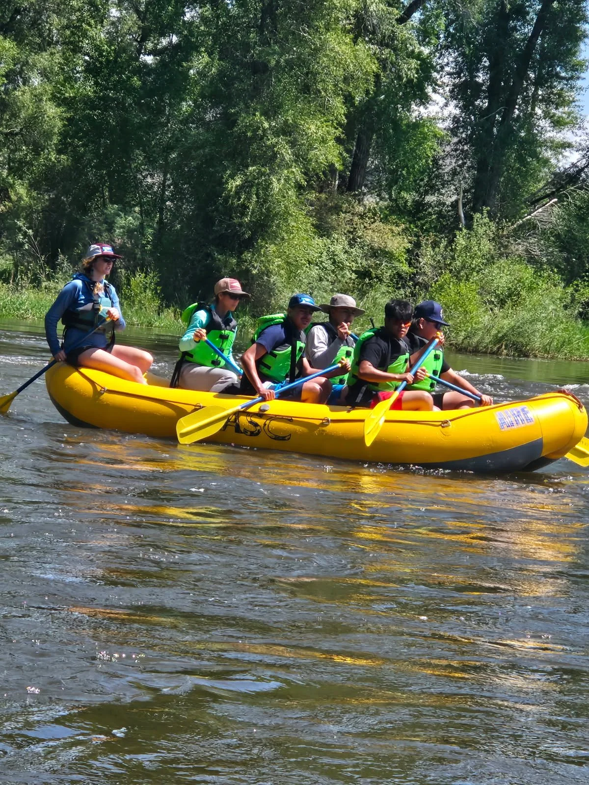 Group of six people in life jackets and hats paddling a yellow inflatable raft on a river surrounded by greenery and trees.