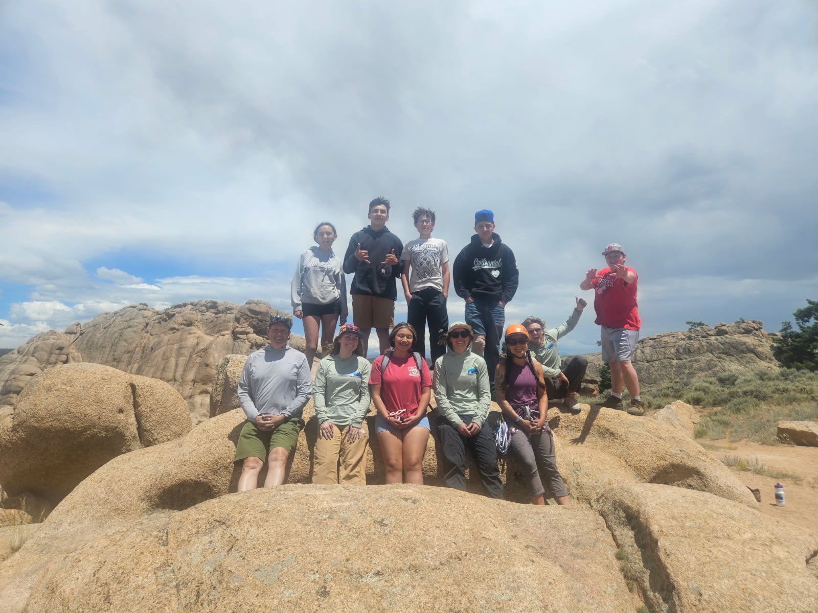 Group of people standing and sitting on large rocks in a rocky outdoor area with a cloudy sky.