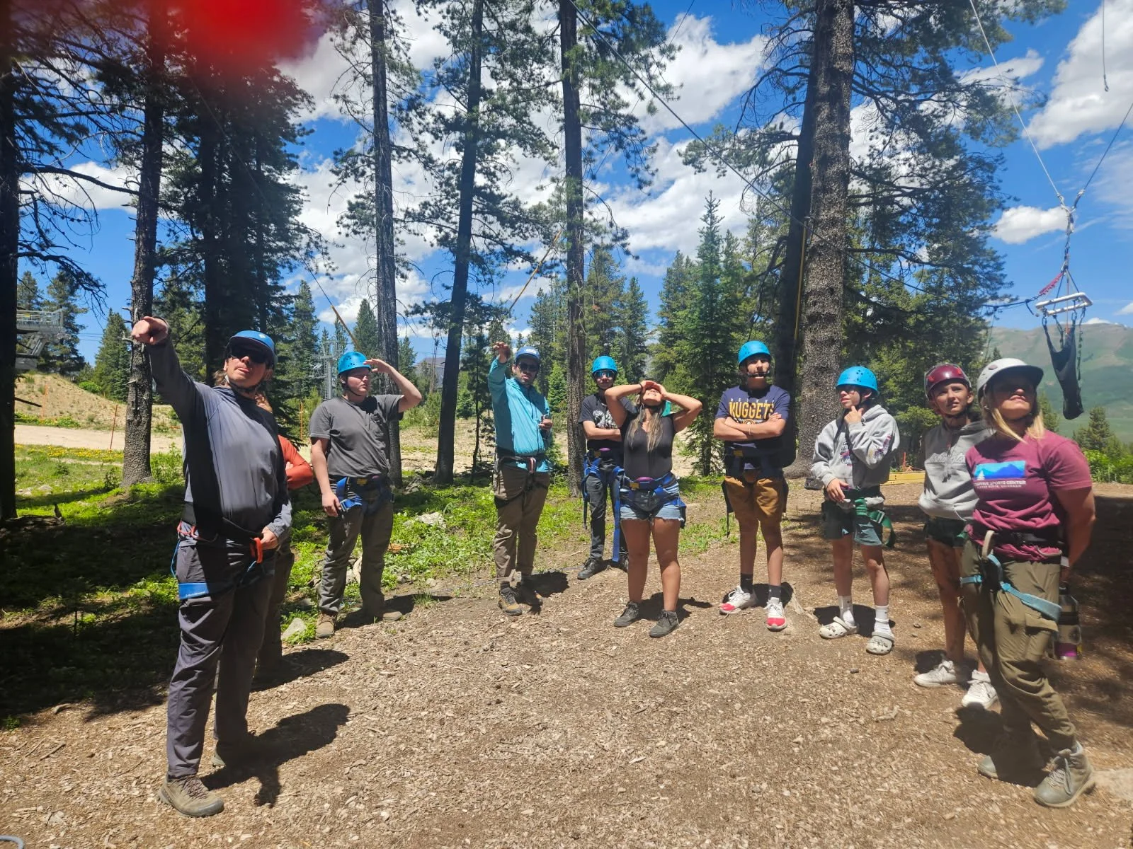 Group of nine people in outdoor gear standing on a dirt trail in a forested mountain setting with trees, blue sky, and clouds.