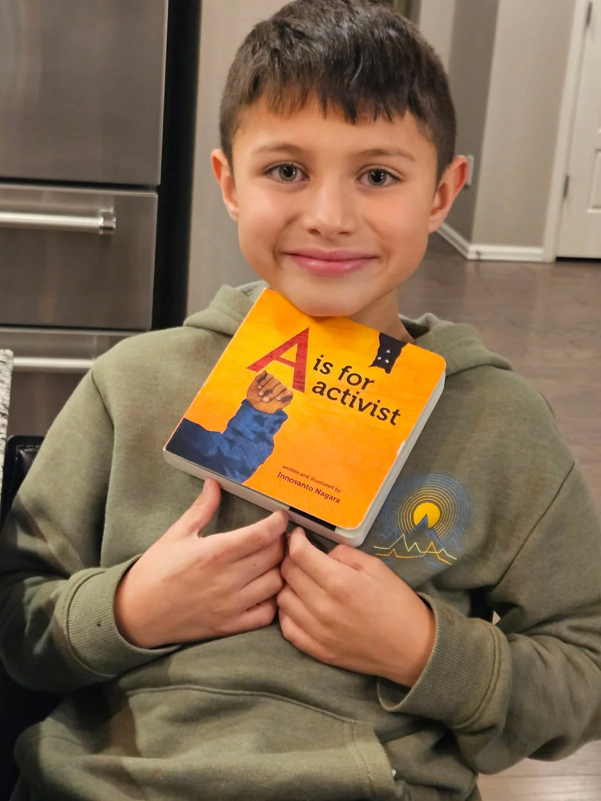 A young boy with brown hair and blue eyes smiling while holding a book titled 'A is for activist' close to his chest, sitting in a kitchen.
