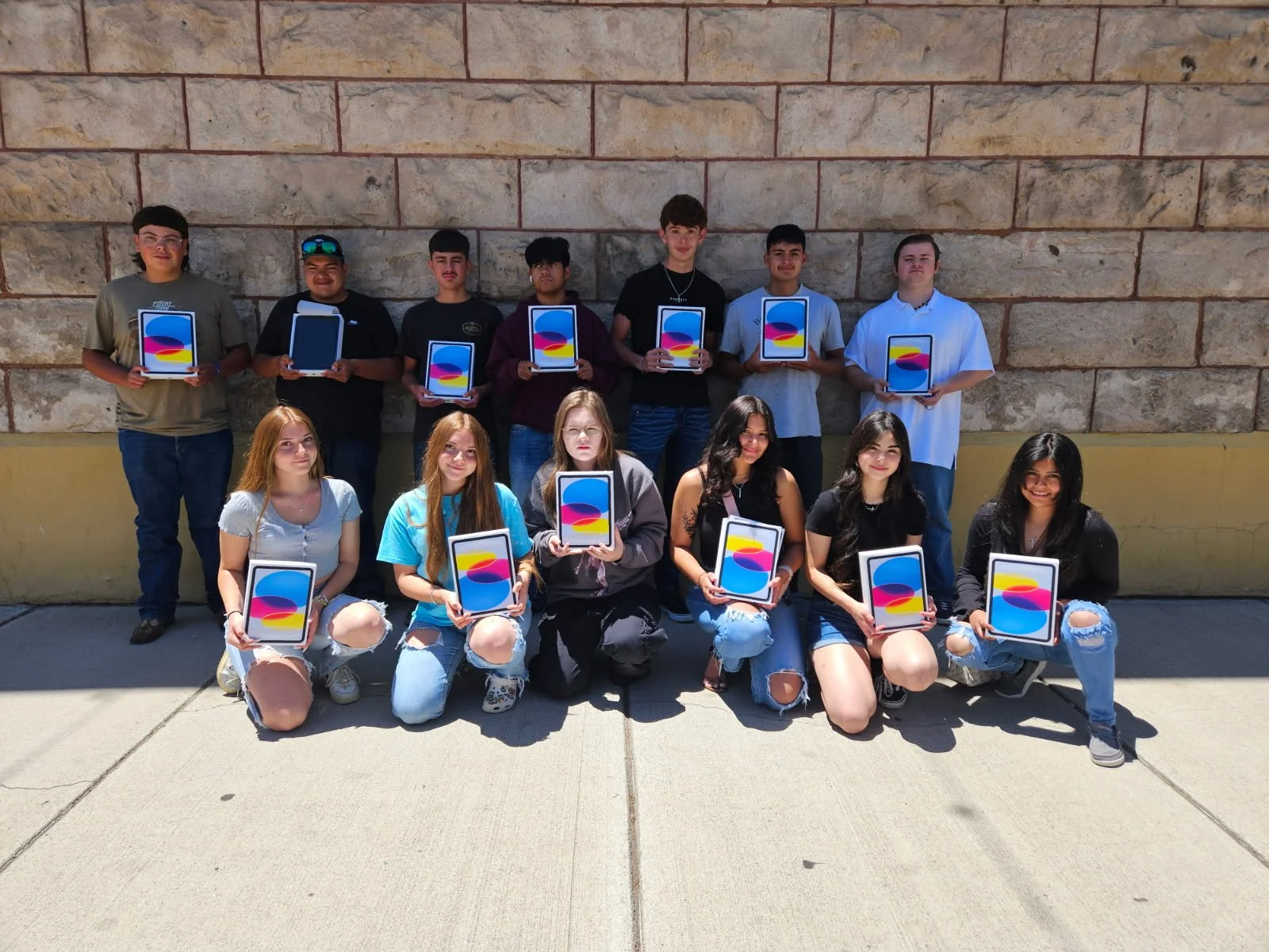 Group of teenagers holding tablets with a colorful abstract background, standing and kneeling outdoors against a stone wall.