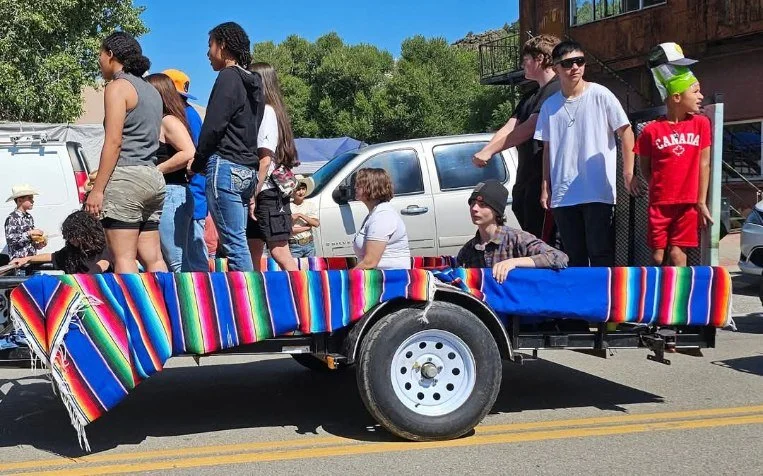 A group of people riding on a colorful, striped trailer during a parade or community event on a sunny day.