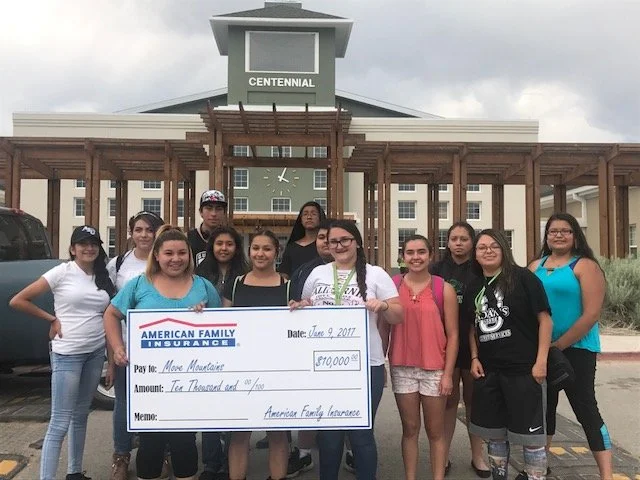 Group of young women standing outdoors in front of a building holding a large check for $10,000 from American Family Insurance, with the date June 9, 2017.