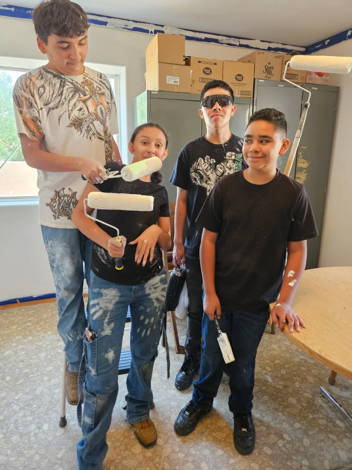 Four children painting a room with white paint, with paint rollers in hand. They are covered in paint splatters and appear to be working on a home renovation project.