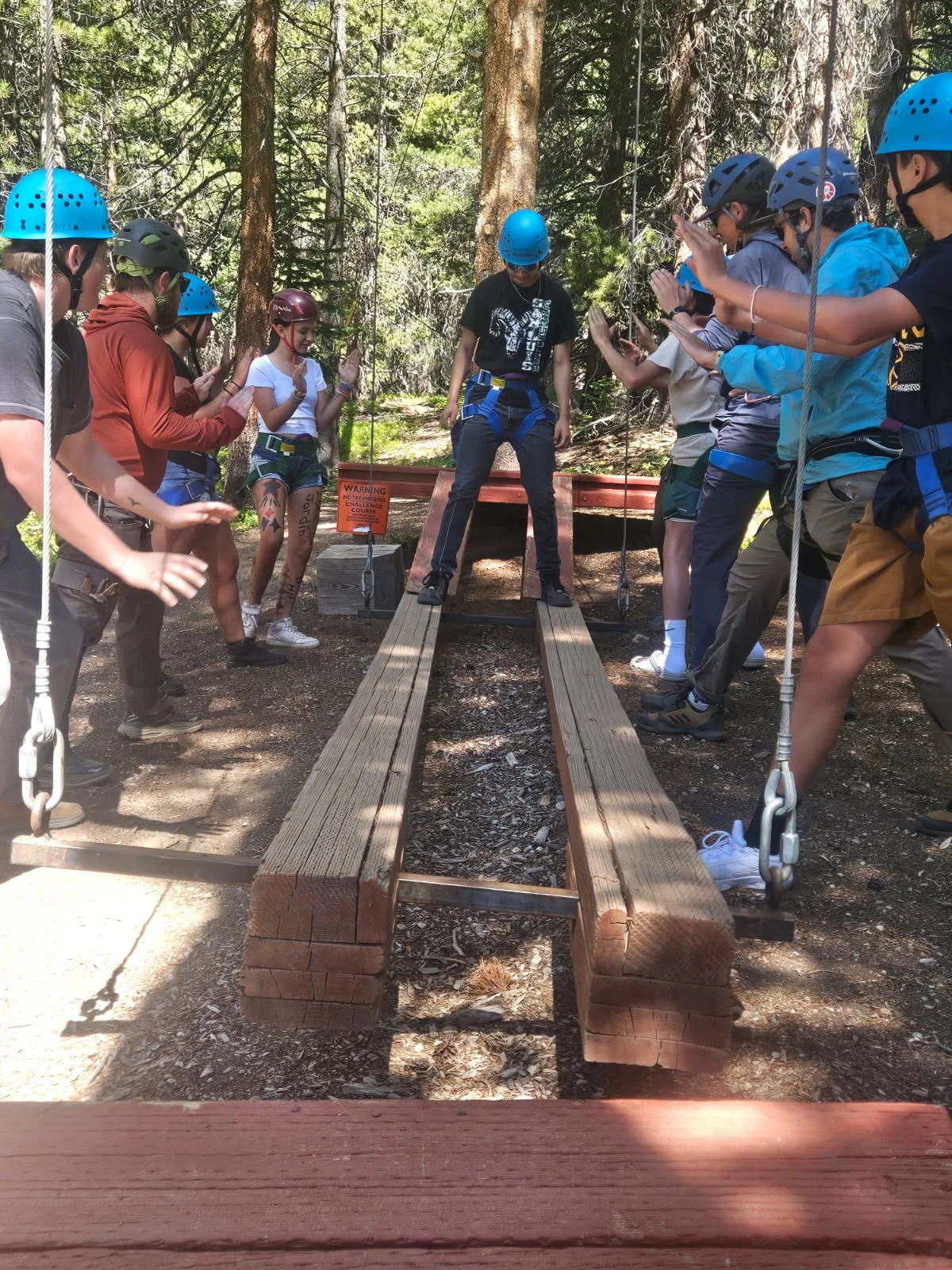 Group of people wearing helmets and harnesses standing and waiting on a wooden bridge in a forest during outdoor adventure activity.