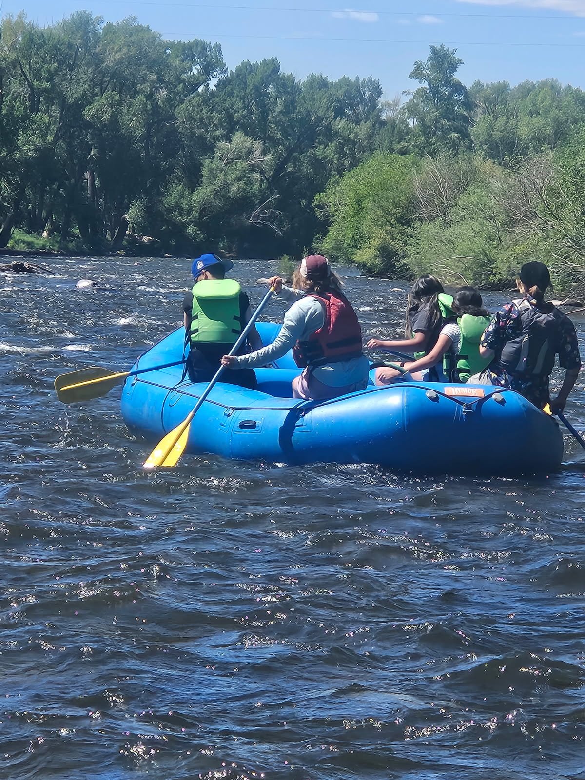 Group of six people in a blue inflatable raft, wearing life jackets, paddling on a river surrounded by green trees on a sunny day.