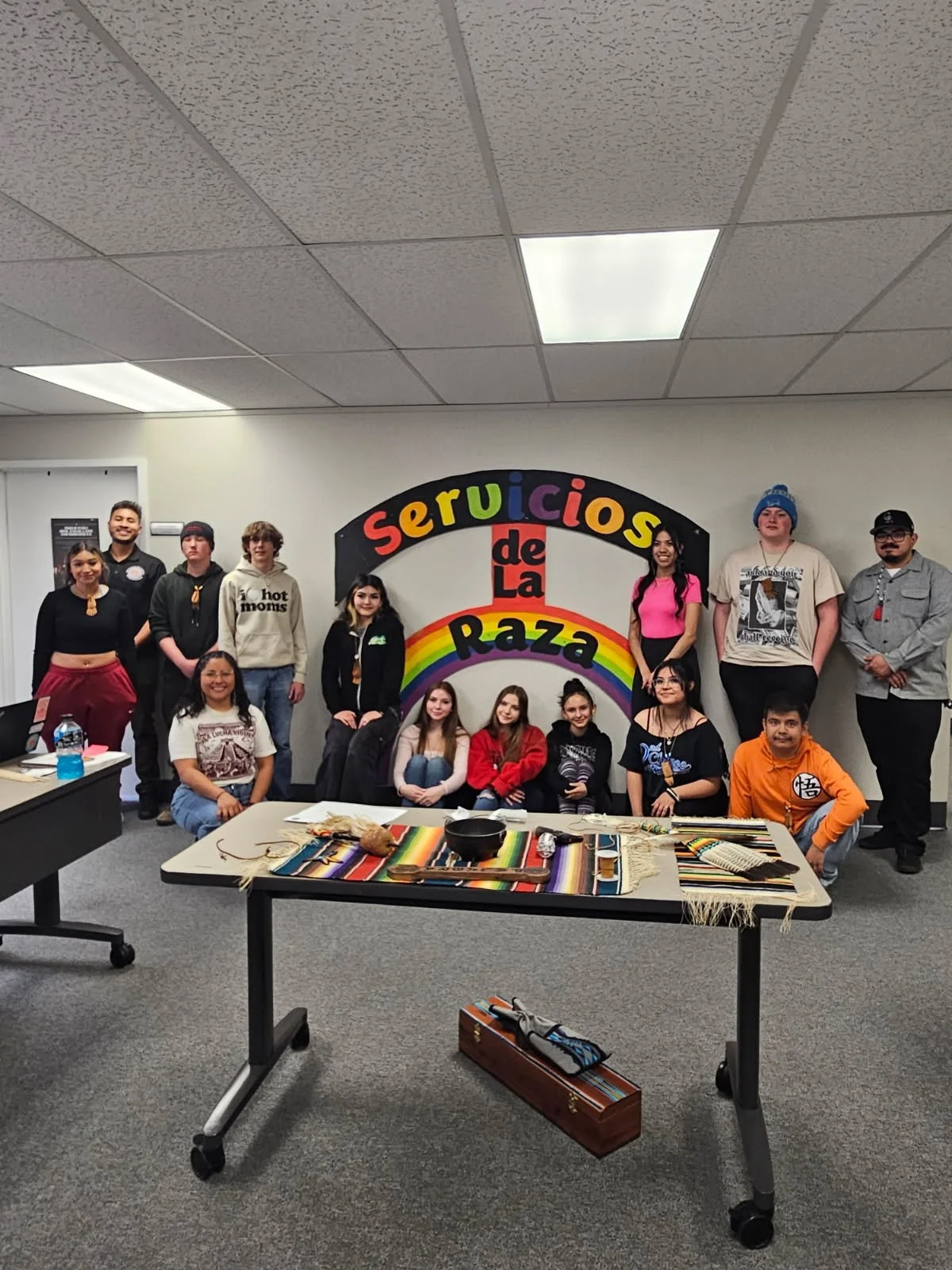 Group of diverse young people gathered in front of a colorful mural with the words 'Servicios de la Raza' and a rainbow. The group is standing and sitting in a room with tables displaying colorful woven textiles and craft supplies.