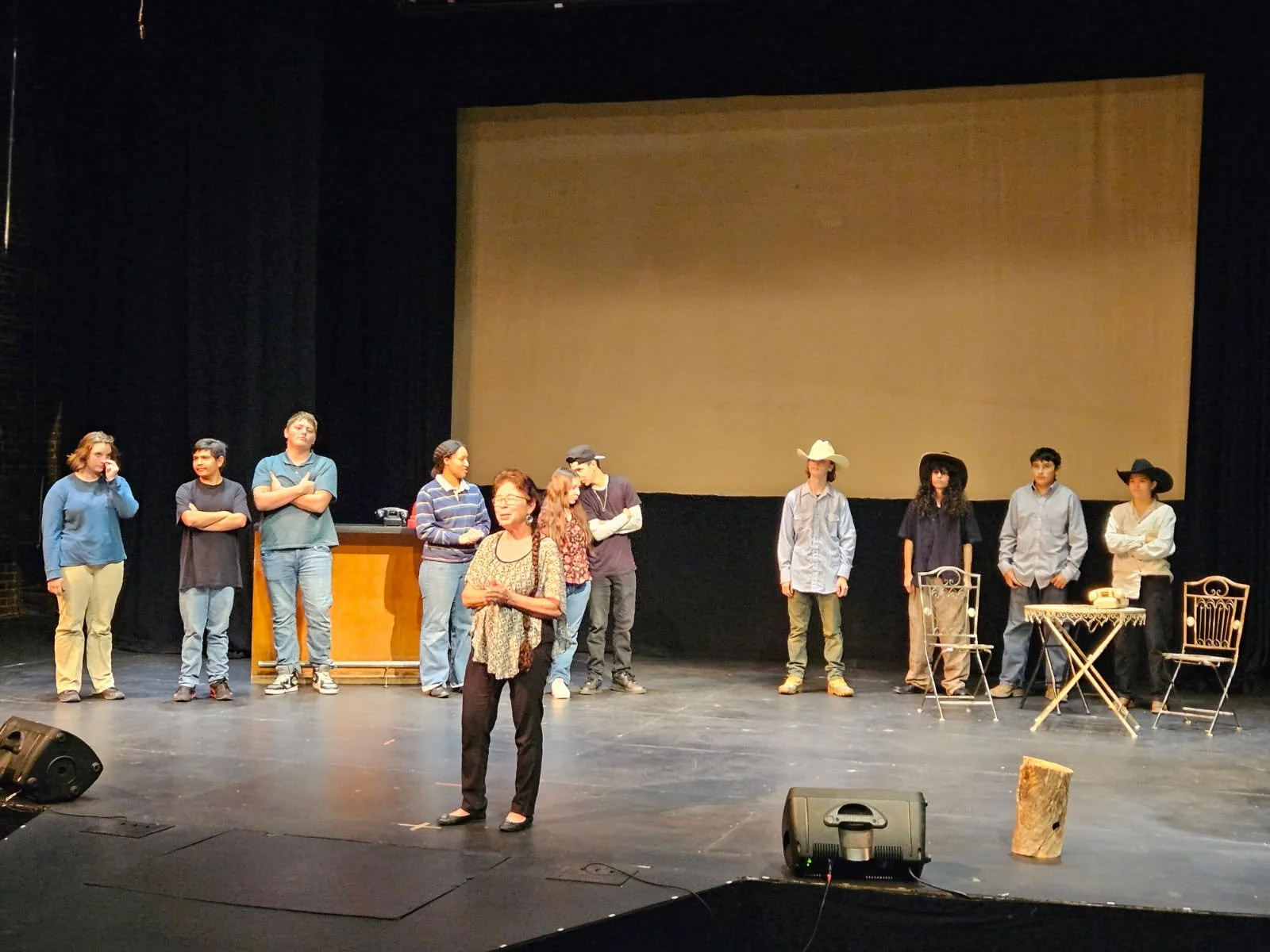 A group of actors on stage during a rehearsal or performance, with some dressed in casual clothing and cowboy hats, and others standing behind a table and chairs. A woman in the foreground appears to be speaking or directing.