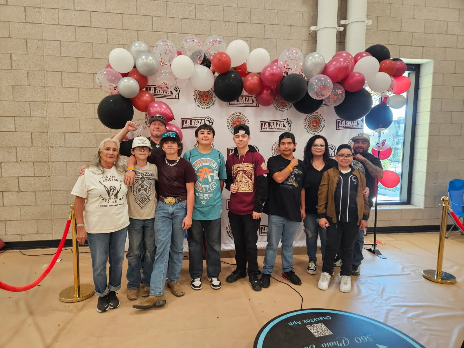 Group of people, including children and adults, posing in front of a backdrop decorated with balloons in black, white, red, pink, and transparent with red confetti, at an indoor event with a red velvet rope barrier.