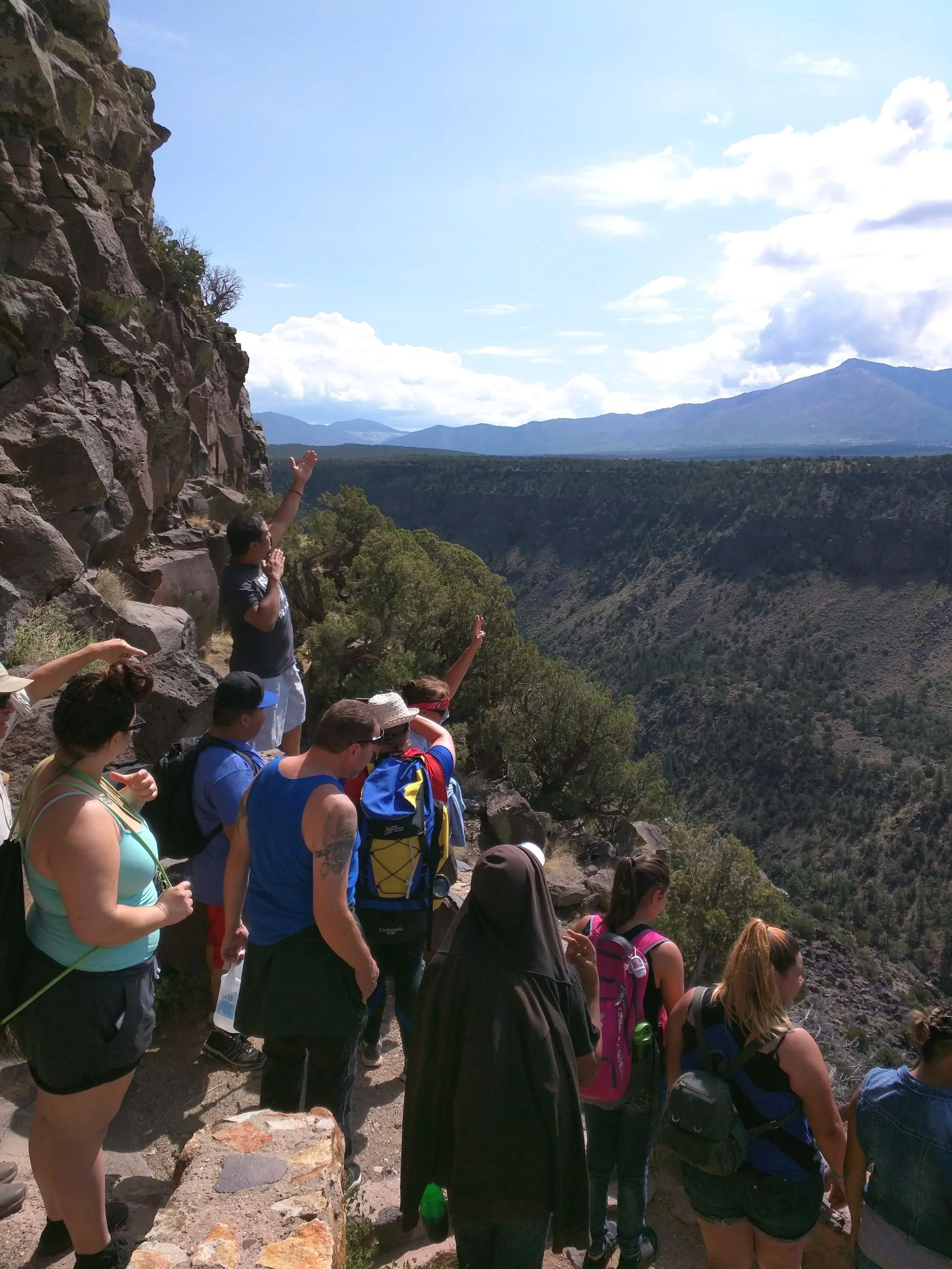 A group of hikers on a trail, pointing towards a distant mountain range under a partly cloudy sky.