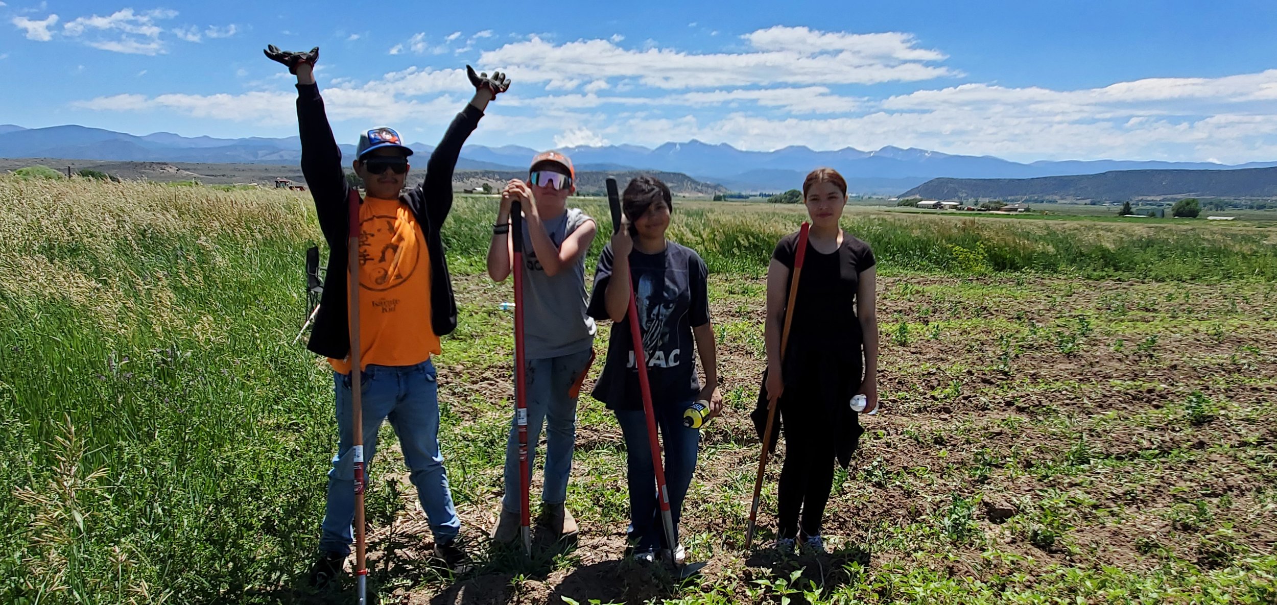 Four children standing in a field with mountains in the background, holding tools and water bottles, under a partly cloudy sky.