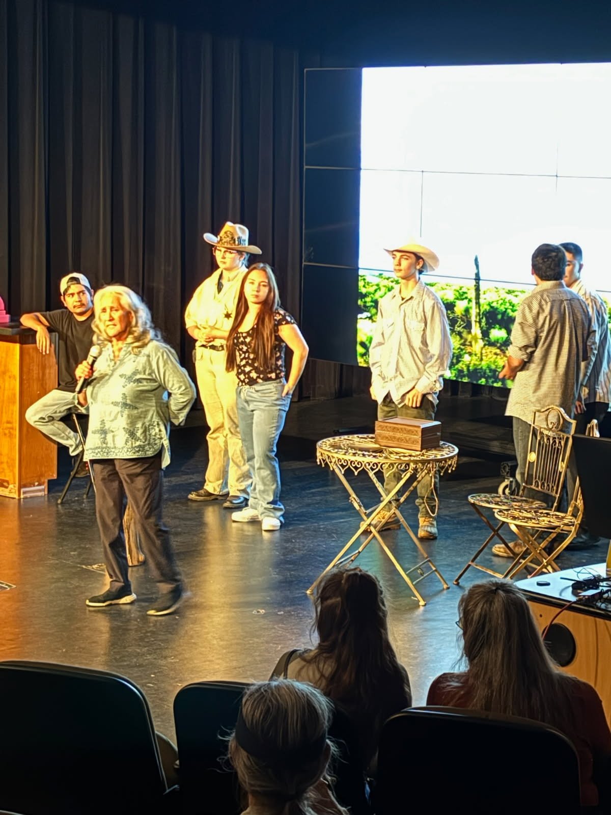 Stage performance with seven people, including a woman with a microphone, a woman in a cowboy hat, and a man dressed as a sheriff, with a large digital screen in the background showing outdoor scenery, and audience seated in front.