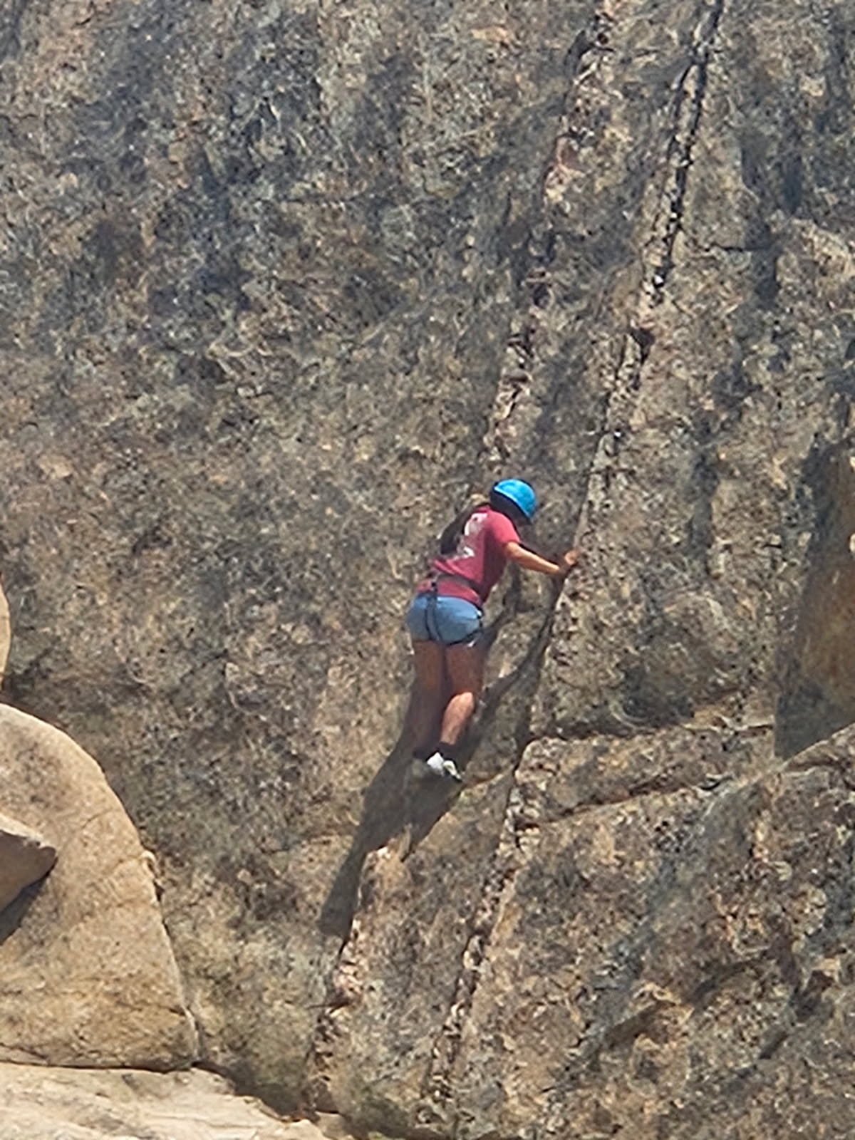Person rock climbing on a steep granite wall wearing a blue helmet, pink shirt, and blue shorts.