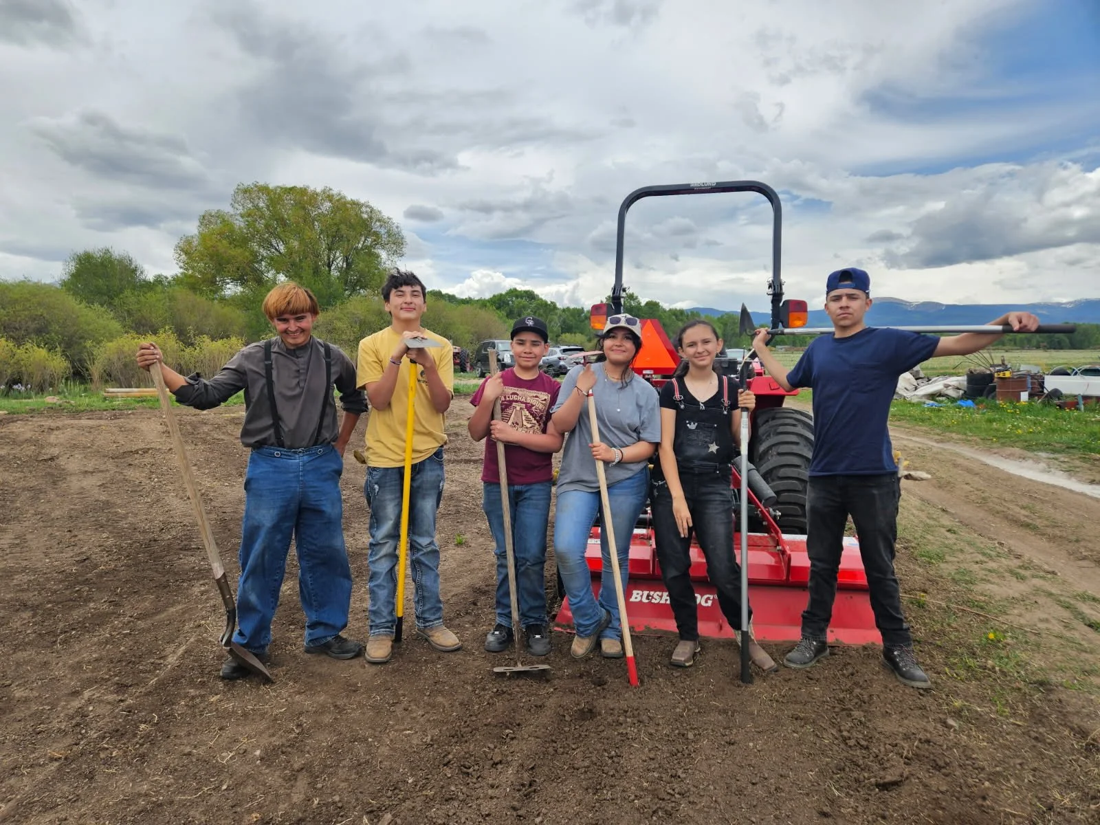 Six children standing on a farm field next to a tractor, holding gardening tools, with trees and cloudy sky in the background.