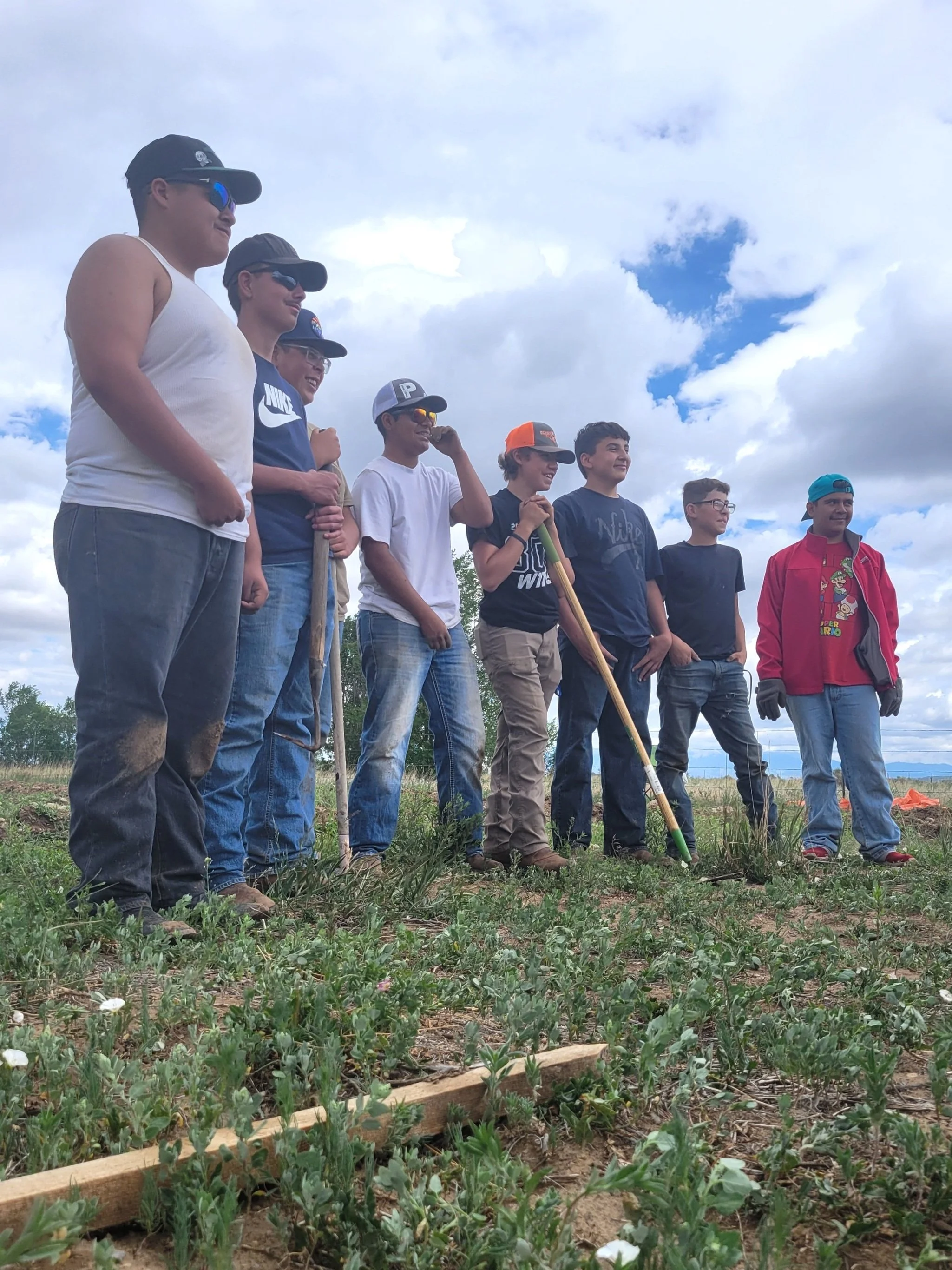 A group of nine young boys standing outdoors on a cloudy day in a field, some holding gardening tools, ready to plant or tend to a garden.
