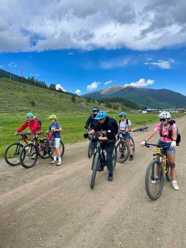 Group of six children with helmets and bikes on a dirt trail near green hills and mountains under a partly cloudy sky.