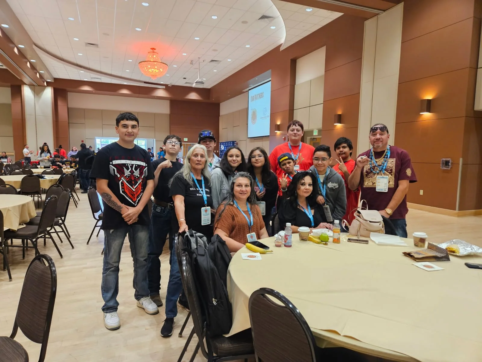 Group of people, including children and adults, gathered around a table in a large conference room with beige walls and a chandelier.