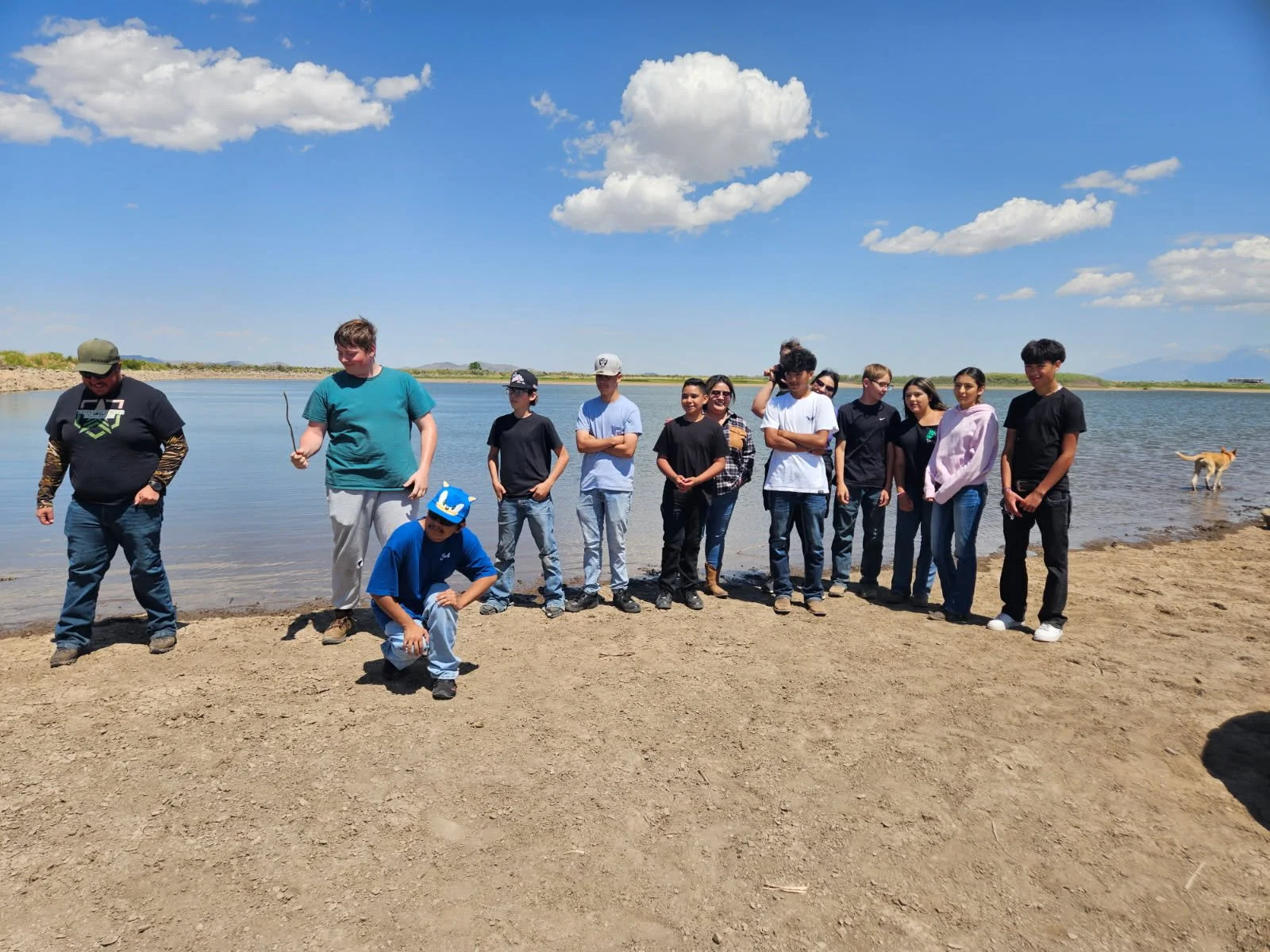 A group of people standing and kneeling on a sandy beach by a body of water on a sunny day with a blue sky and scattered clouds.