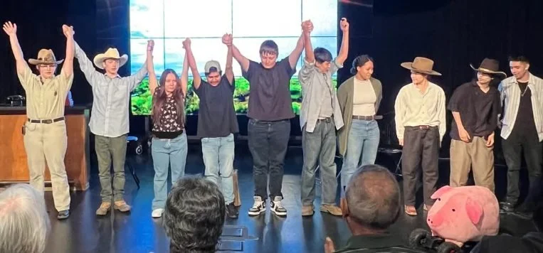 Group of nine people on stage holding hands up in celebration, some wearing cowboy hats, with an audience watching and a large digital screen displaying a nature scene in the background.
