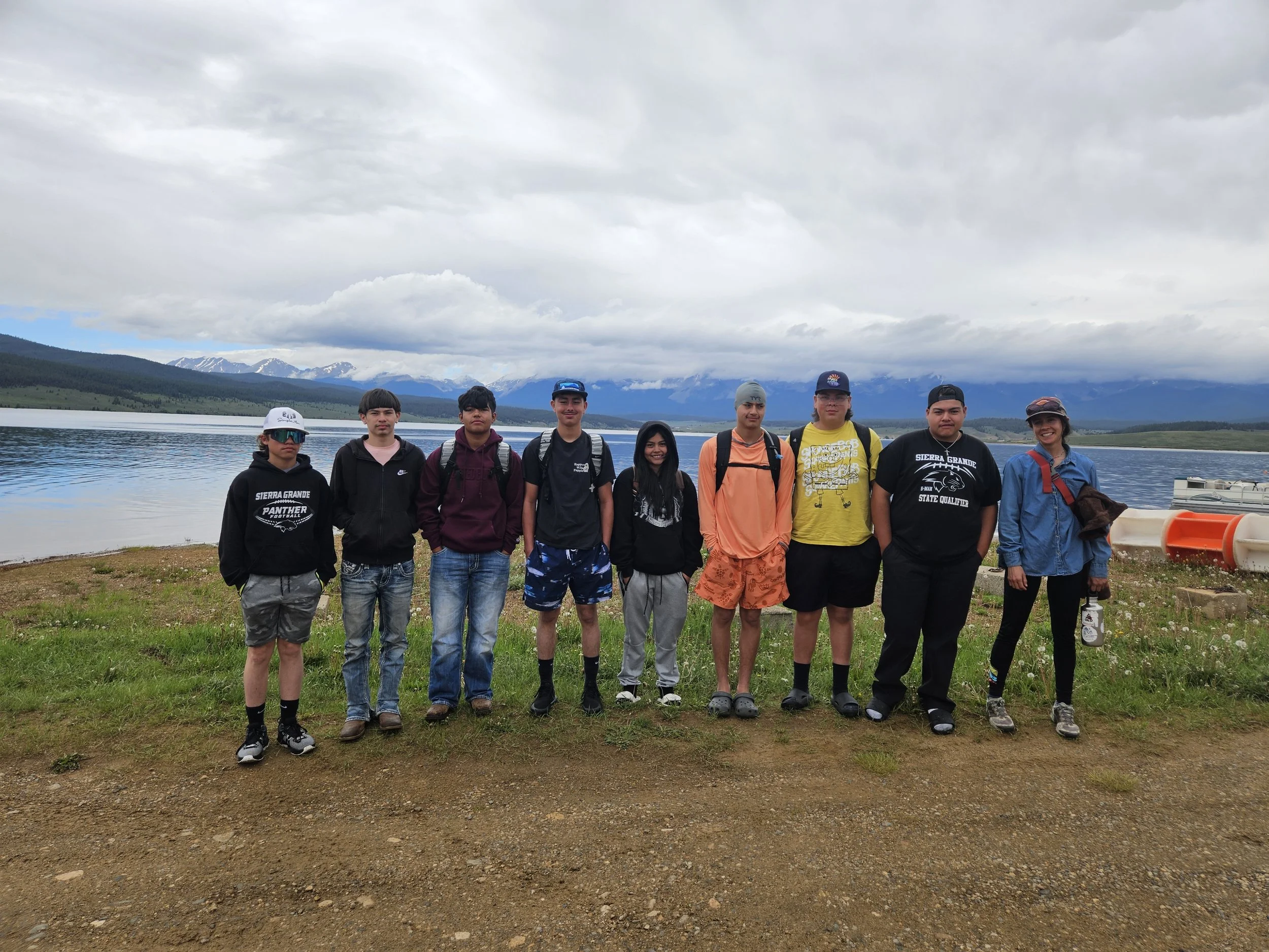 Group of nine young people standing by a lake with mountains and cloudy sky in the background.