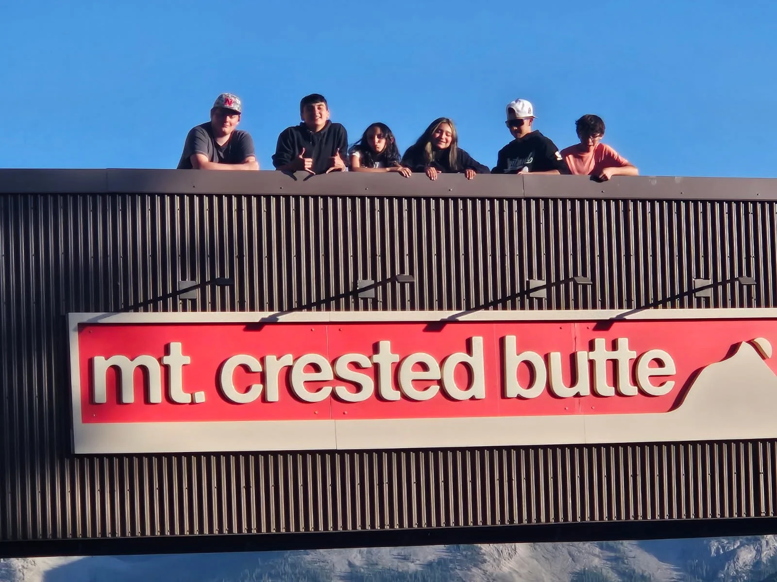 Six people standing on a rooftop above a sign that reads 'mt. crested butte' with snow-capped mountains in the background.