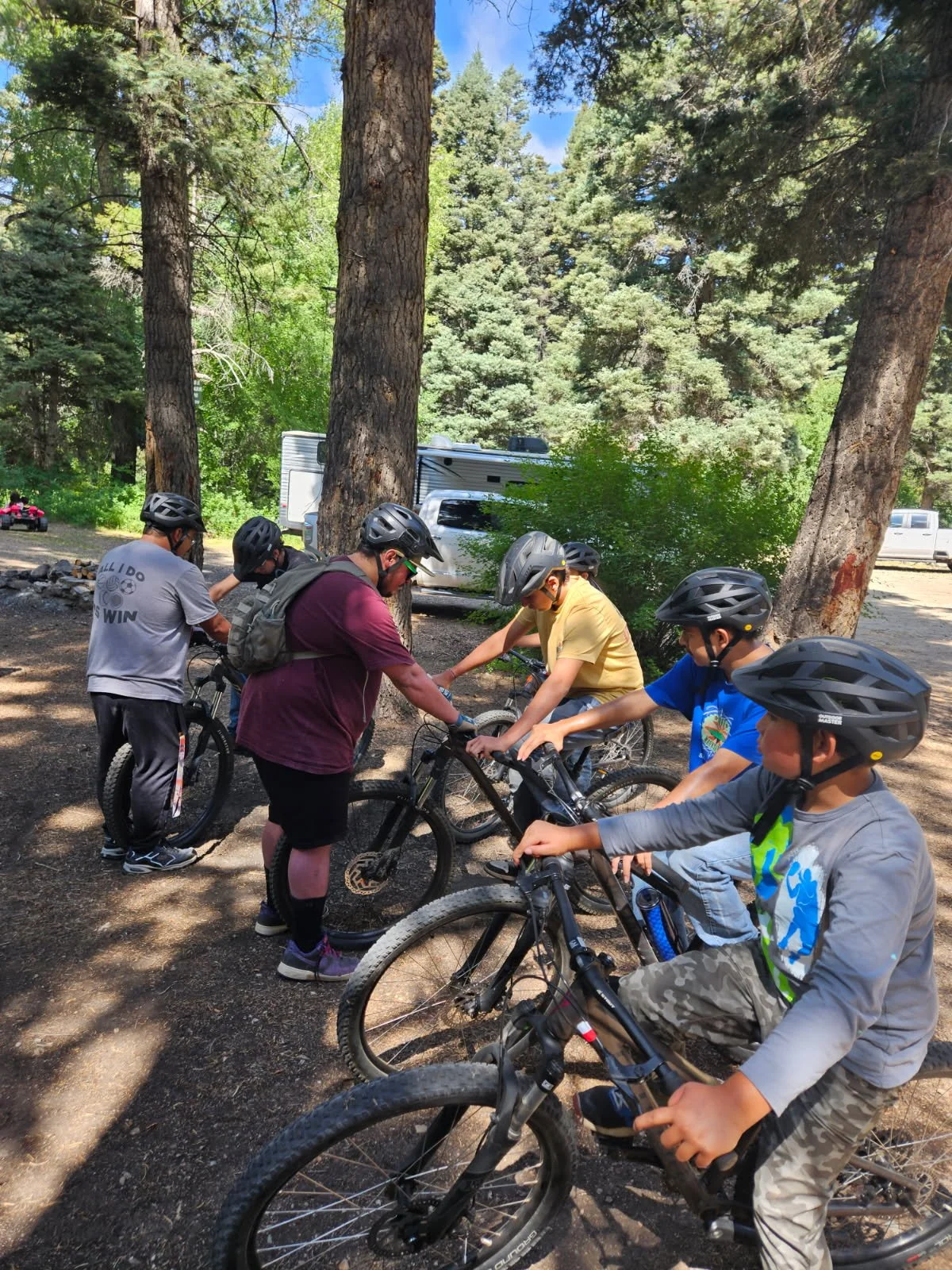 Group of children and a teenager wearing helmets preparing to ride bicycles in a wooded area with tall trees, with a campsite and vehicles in the background on a sunny day.