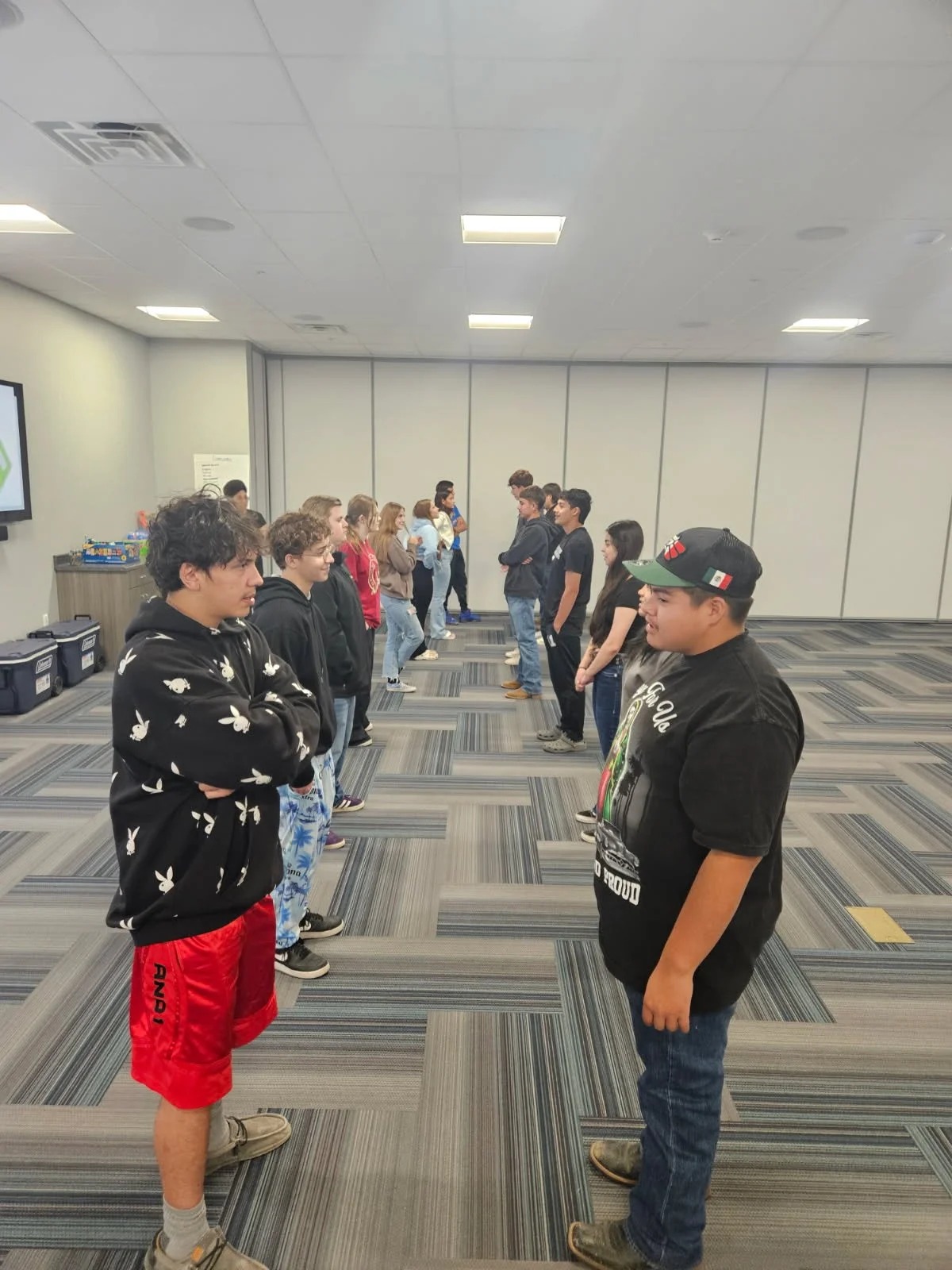 Group of young people standing face-to-face in two lines in a spacious room with gray carpet and white walls, possibly participating in an activity or game.