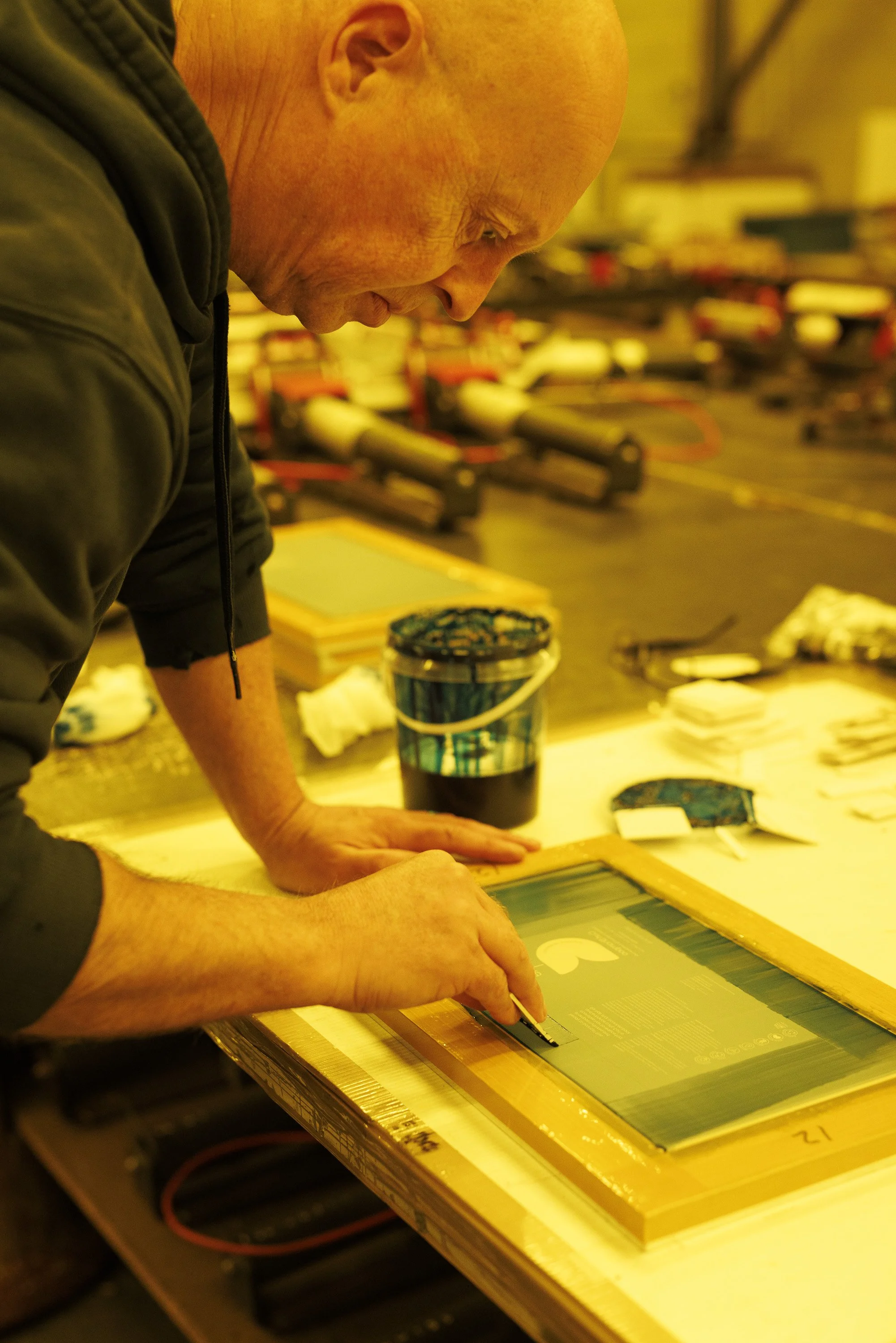 A man with a shaved head working on an art project, leaning over a table with art supplies, with colorful printmaking tools and a screen in front of him.