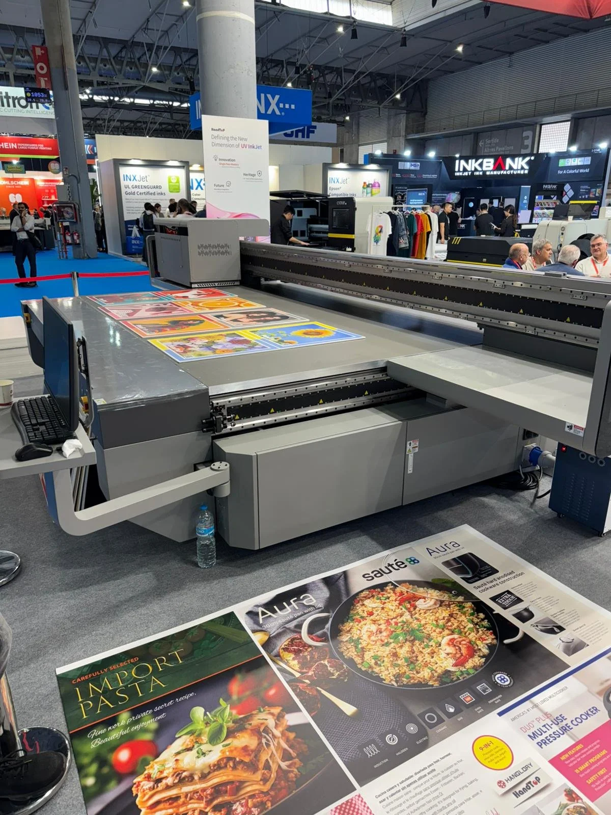 A large industrial printer printing colorful posters or advertisements at a trade show or exhibition. In the background, booths display signage for ink and printing companies, and people are walking around and examining the exhibits.