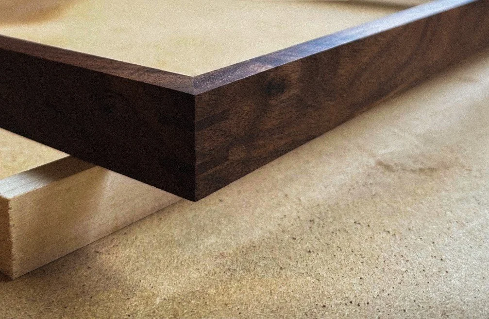 Close-up of a dark wooden frame resting on a piece of lighter wood on a beige textured surface.