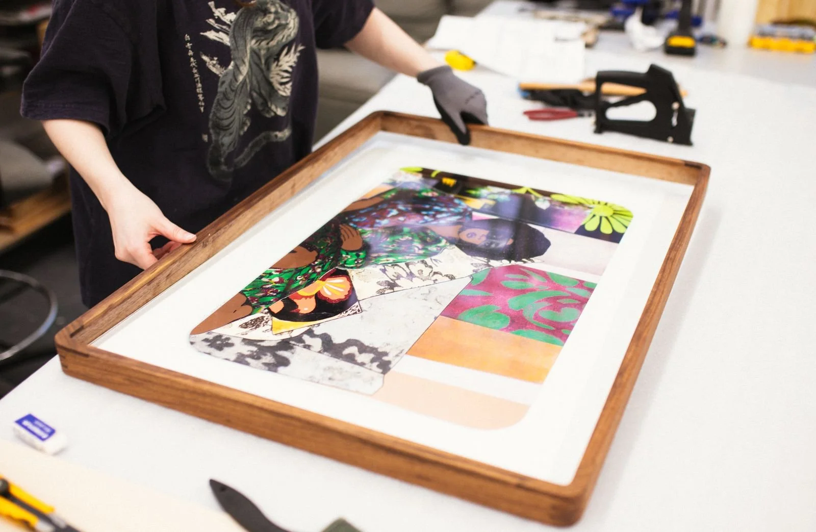 Person holding a framed artwork with colorful collage or textile pieces on a white work table.