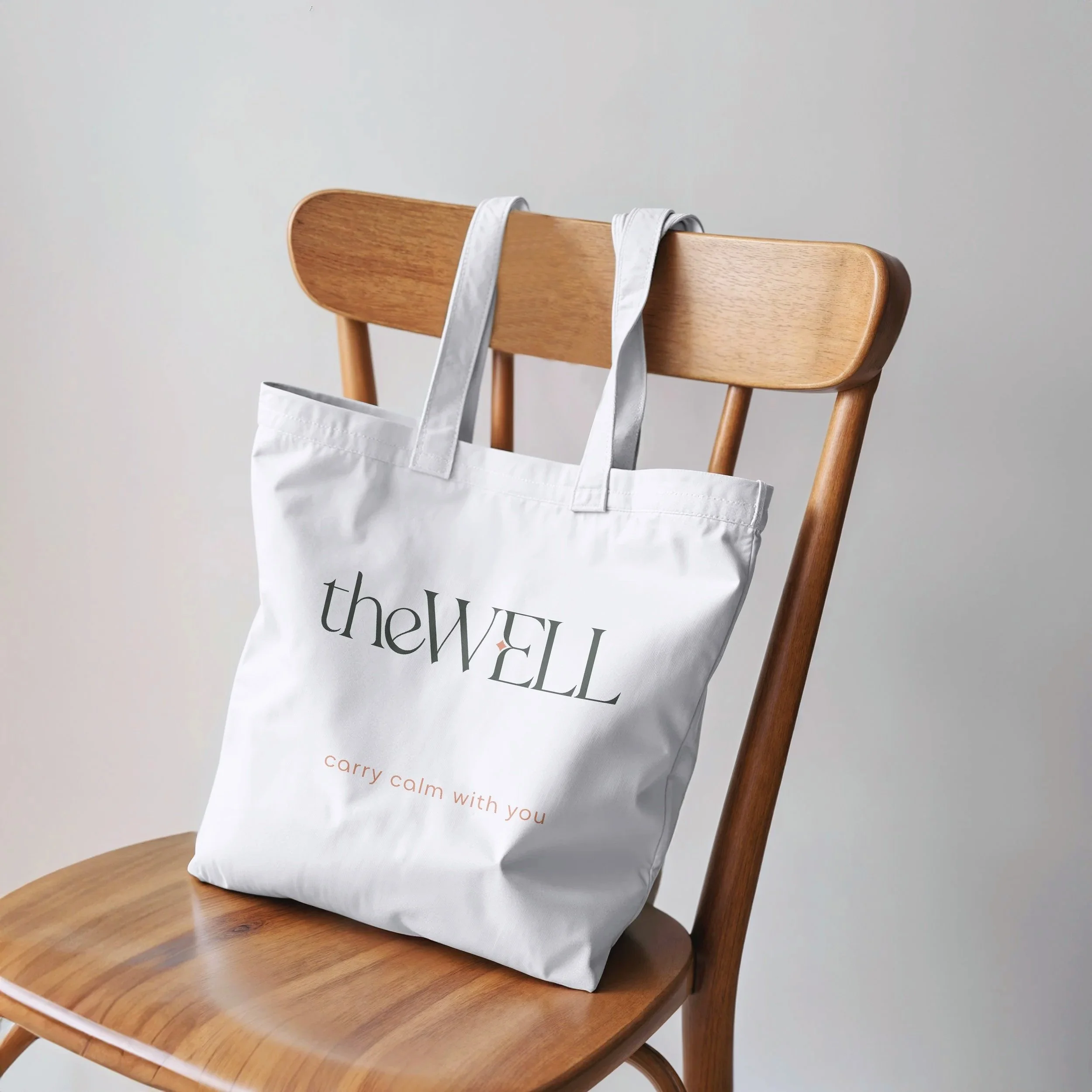White tote bag with the text 'theWELL' and 'carry calm with you' hanging on the back of a wooden chair.