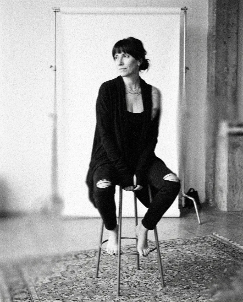 A woman with short hair, wearing a dark clothing, sitting on a tall stool with her feet off the ground, in front of a plain white backdrop, in an industrial-style room.