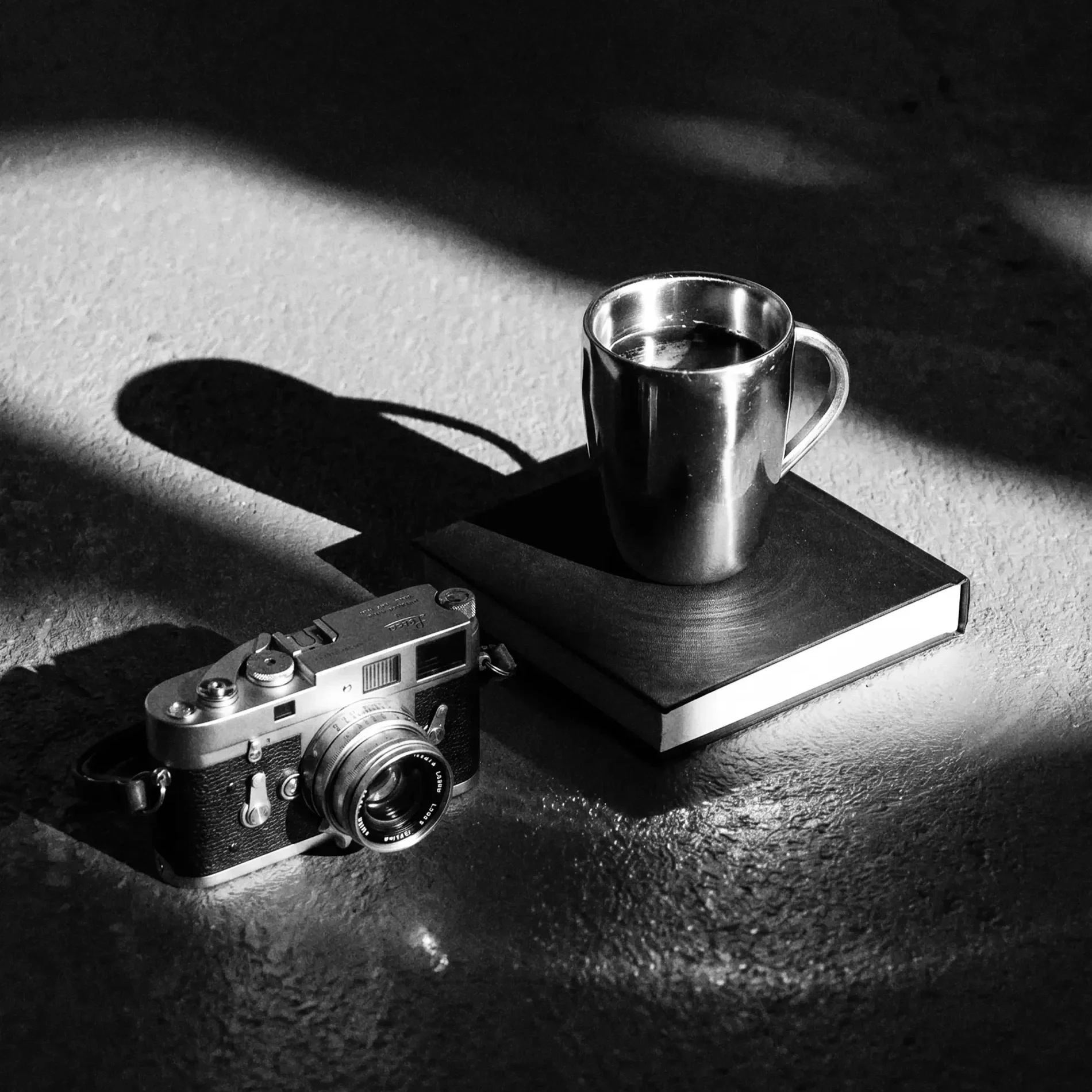 Black and white photo of a metallic camera and a cup of coffee on a textured surface, with shadows cast on the surface.